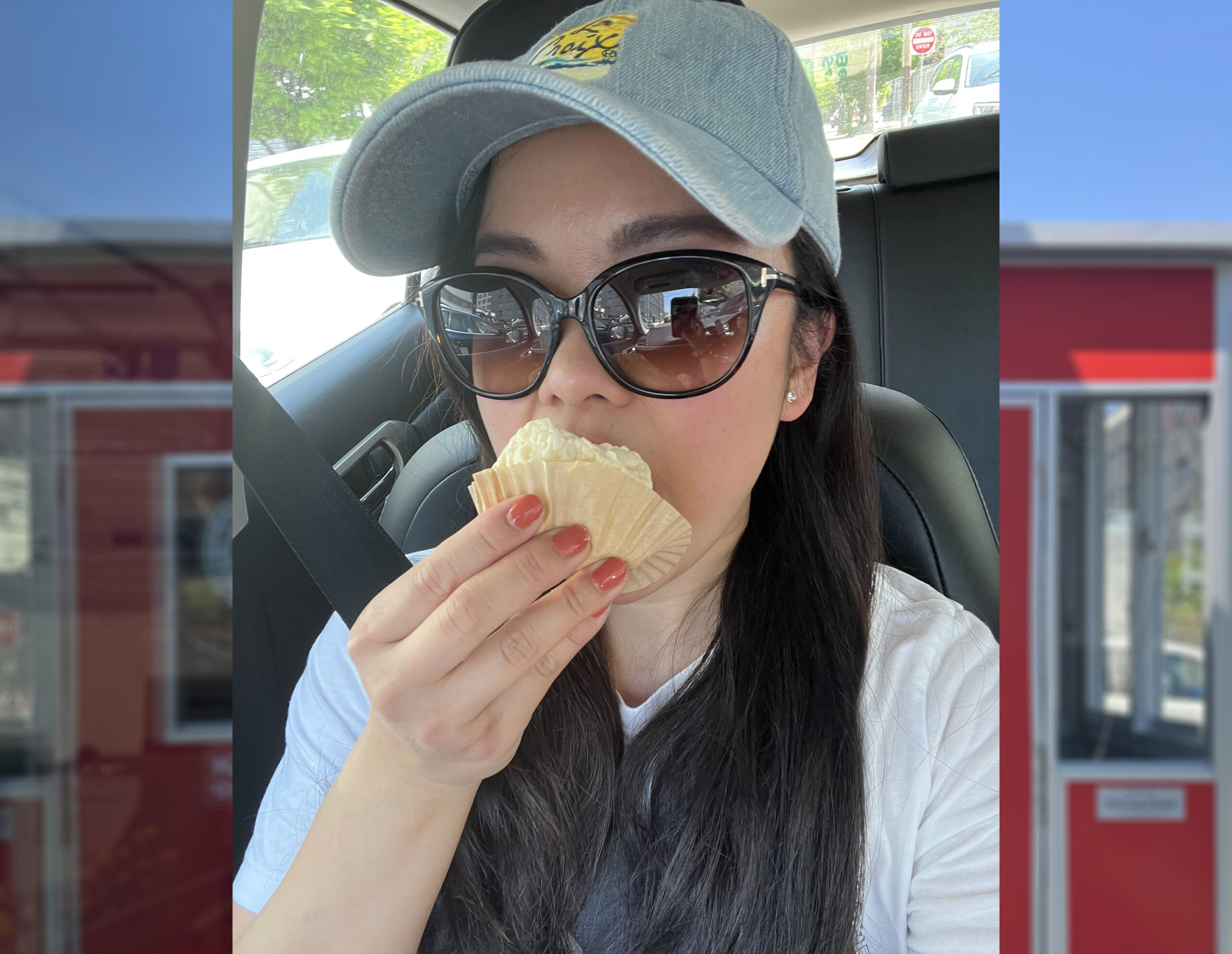 A woman in a car, wearing a baseball cap and sunglasses, eating a cupcake.