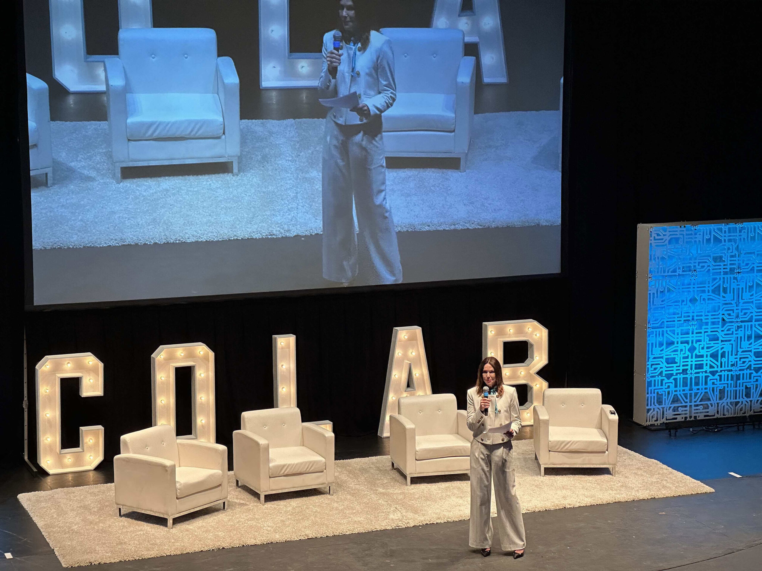 A woman stands on a stage speaking into a microphone. Behind her is a large screen displaying her image, and the stage is set with white chairs and large illuminated letters.