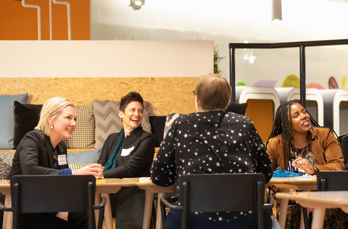 Four people are seated around a table, engaged in a lively conversation. Three of them are smiling or laughing.