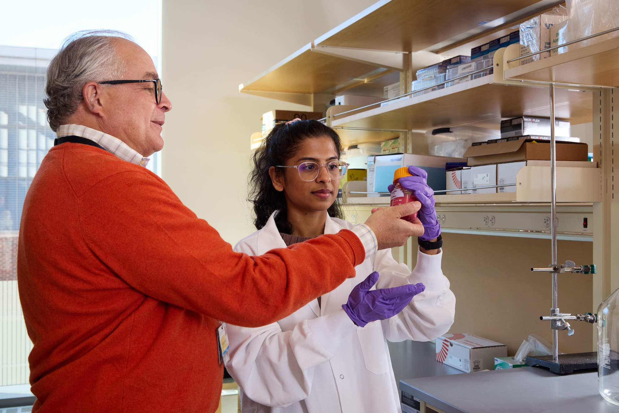 A man in an orange sweater and a woman in a lab coat and gloves examine a container together in a laboratory setting.