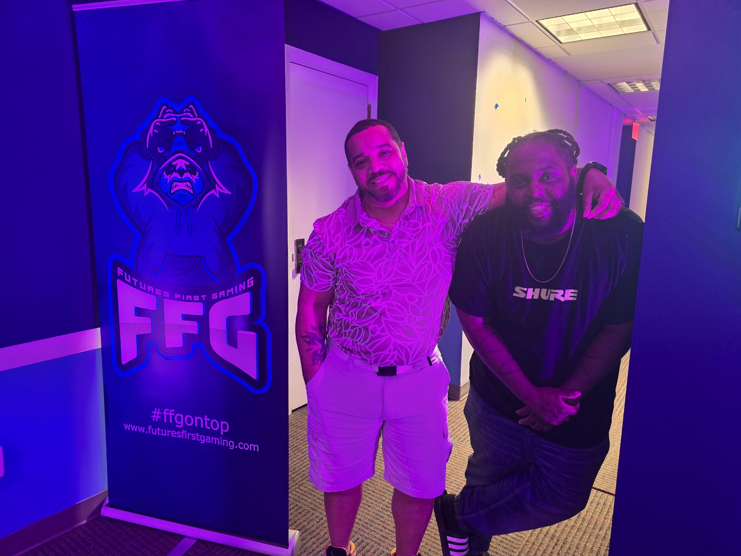 Two individuals stand side by side in a dimly lit room with blue lighting, next to a "Future's First Gaming" banner. One person wears a patterned shirt; the other wears a black "Shure" shirt.