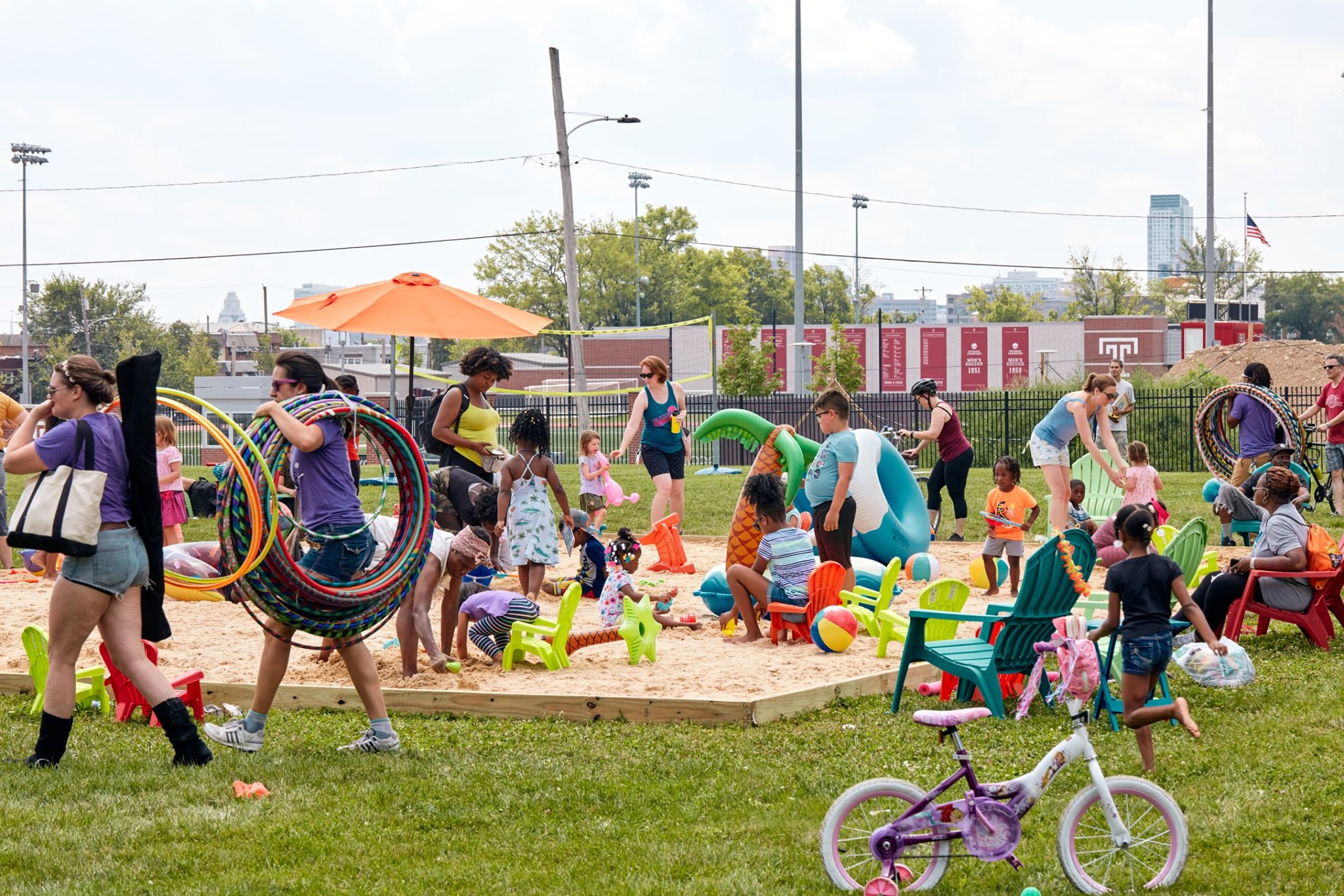 A lively outdoor children's event with various activities, including a sandpit and hula hoops. Adults supervise while kids play. Brightly colored toys and chairs scattered around.