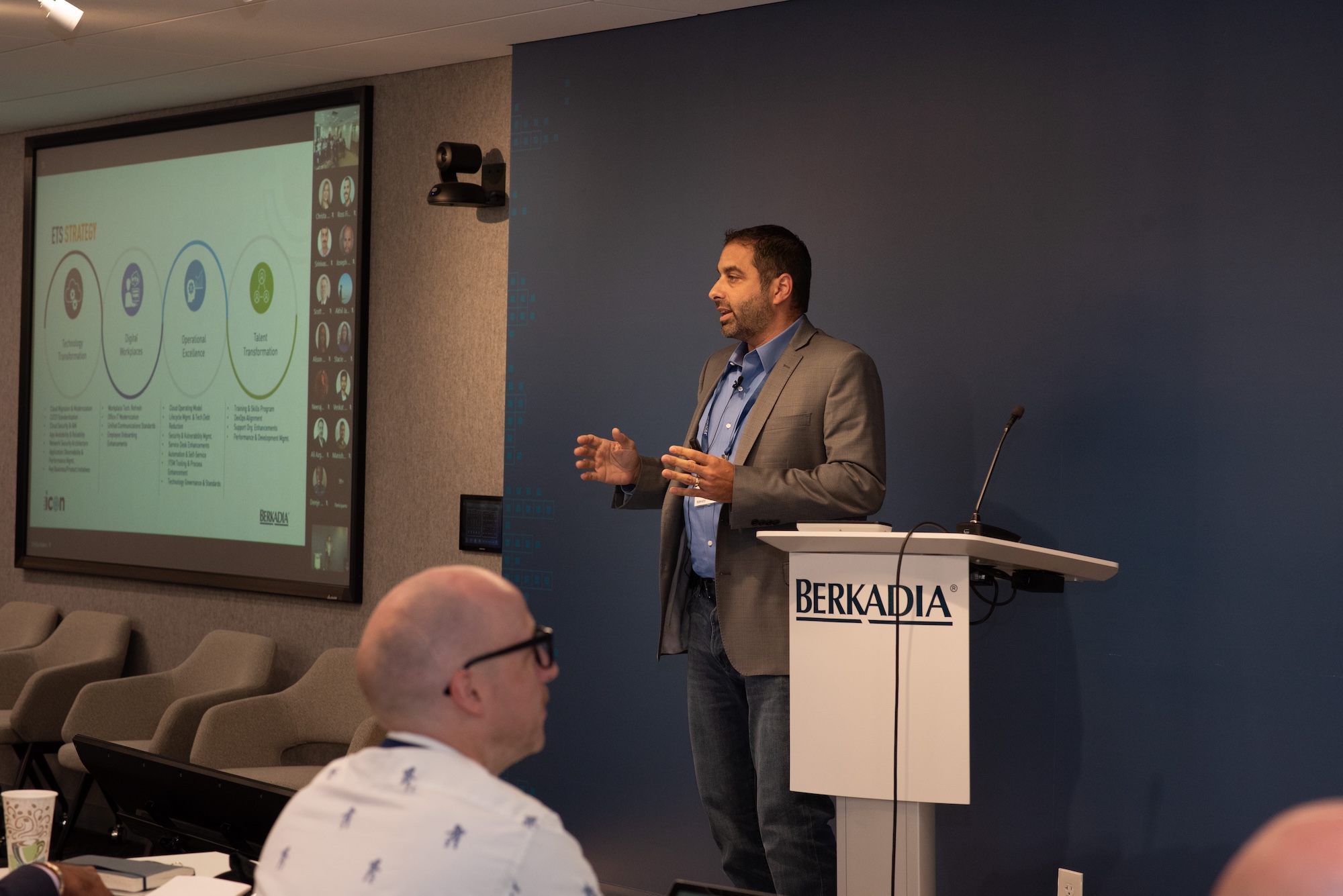 A man standing at the front of a conference room presenting to an audience. He stands next to a podium with the Berkadia logo on it.