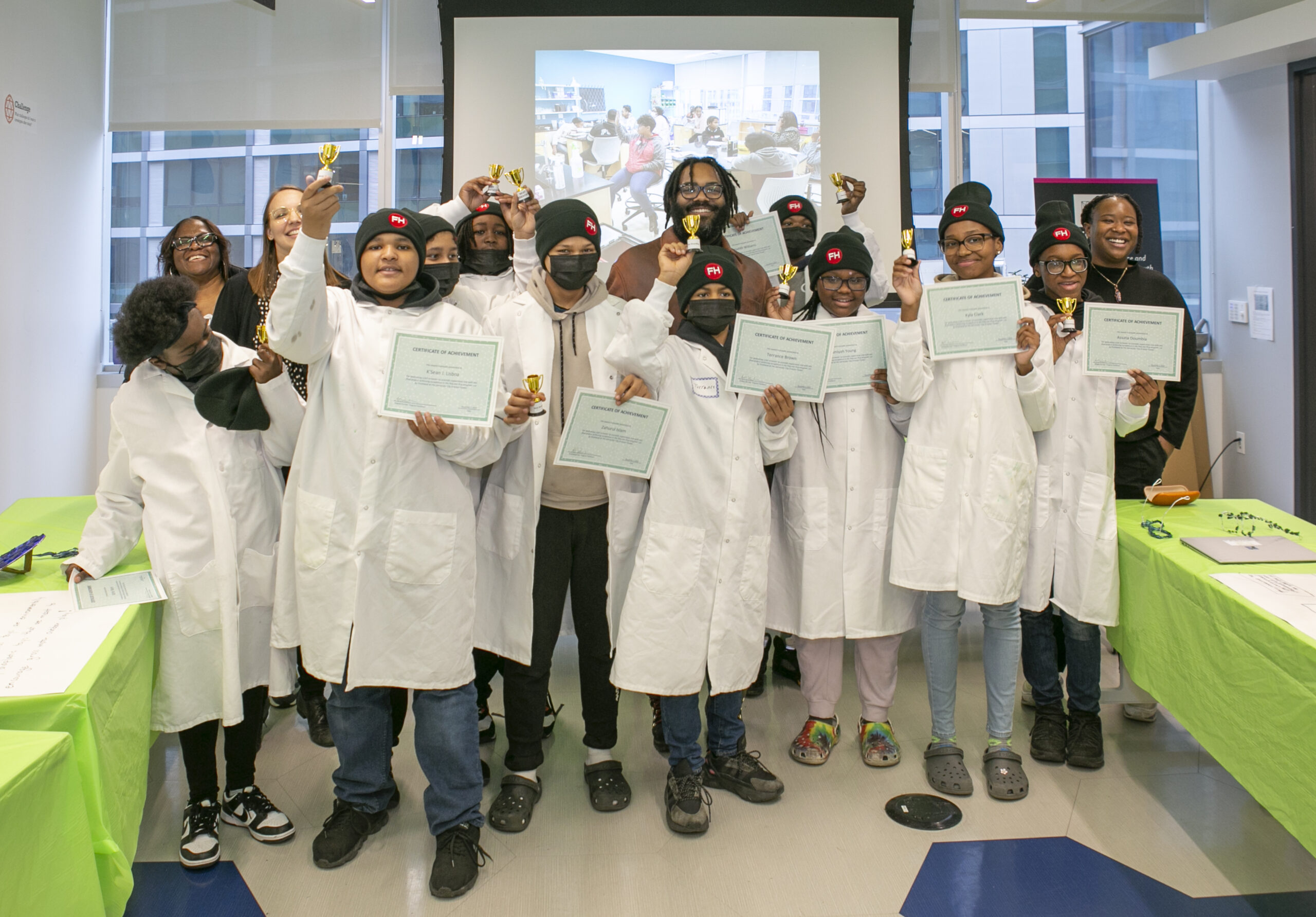 A group of children in lab coats and hats stand together holding certificates and small trophies, smiling at the camera. An adult stands behind them, with a screen displaying a classroom photo in the background.