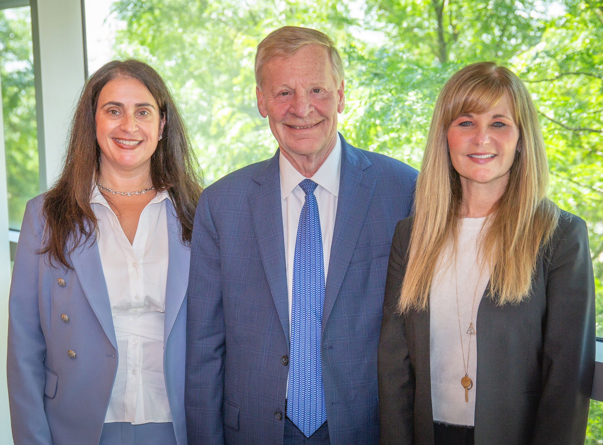 Three white people are standing indoors, smiling at the camera. The man in the center is wearing a blue suit and tie, while the two women on either side are dressed in business attire. A window with greenery is behind them.
