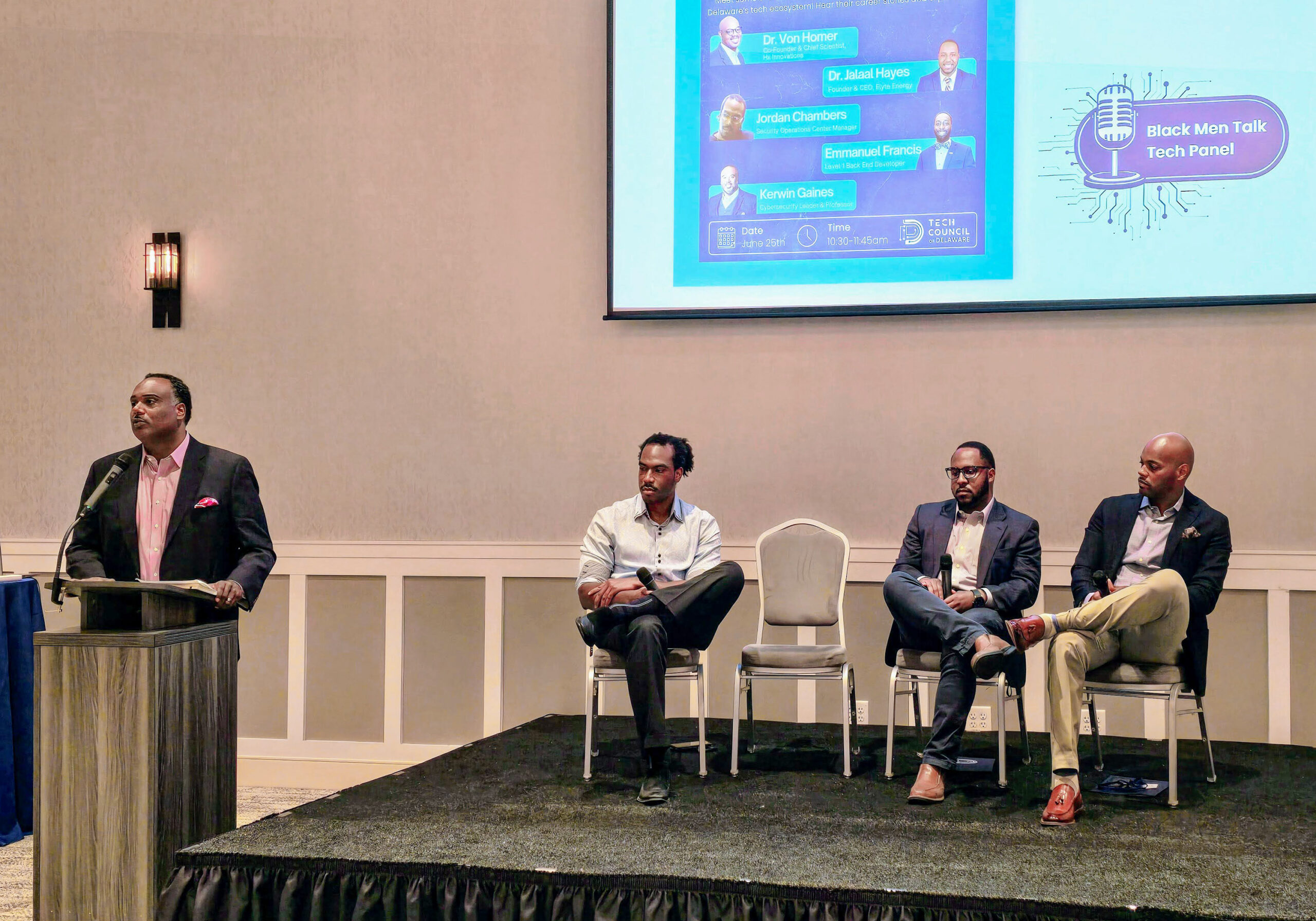 Four Black men are in a conference room. One is speaking at a podium, and three are seated on a stage. A presentation slide behind them reads "Black Men Talk Tech Panel.