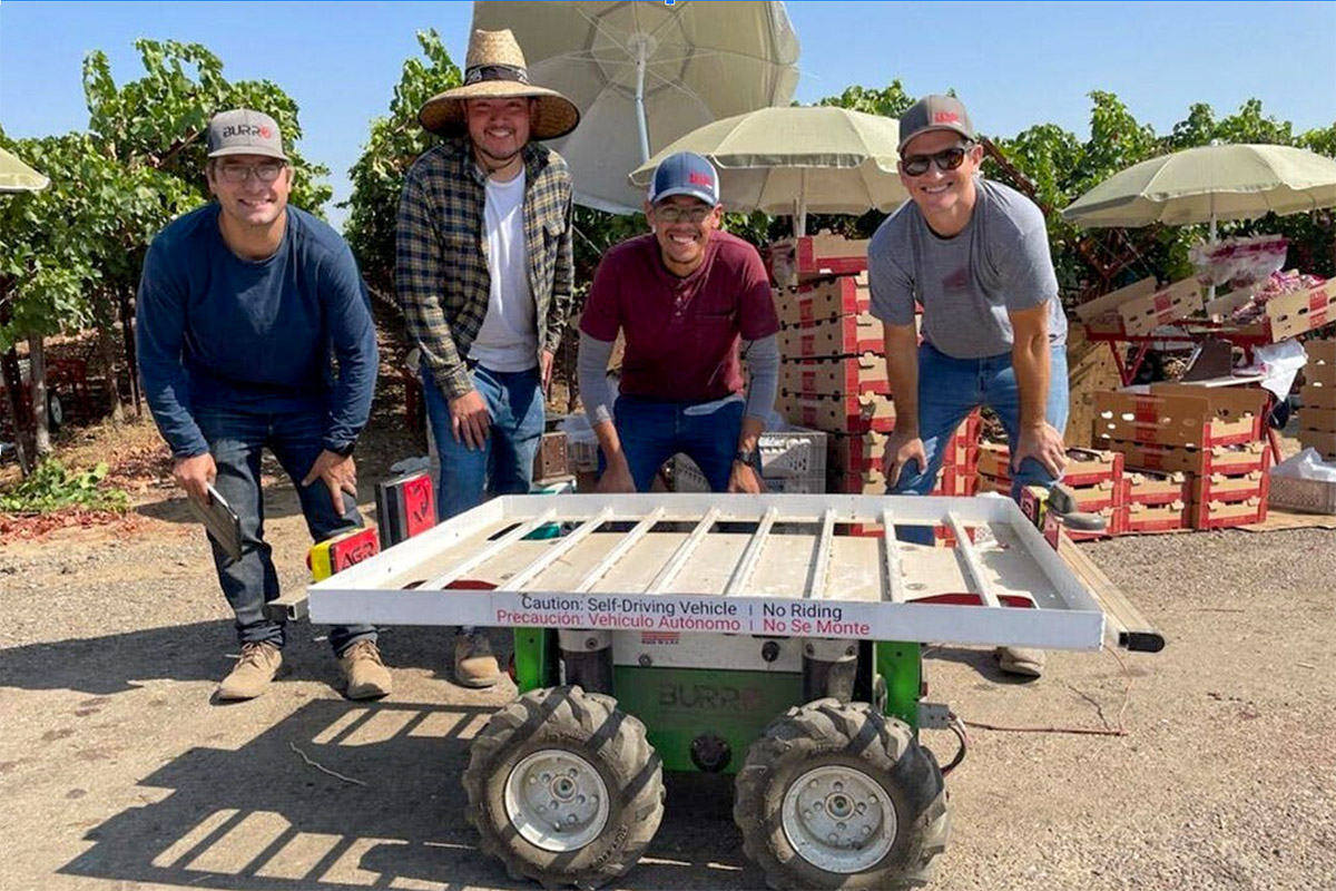 Four people stand behind a small, self-driving vehicle labeled in a vineyard setting. They are smiling, and the vineyard stretches out behind them under a sunny sky.