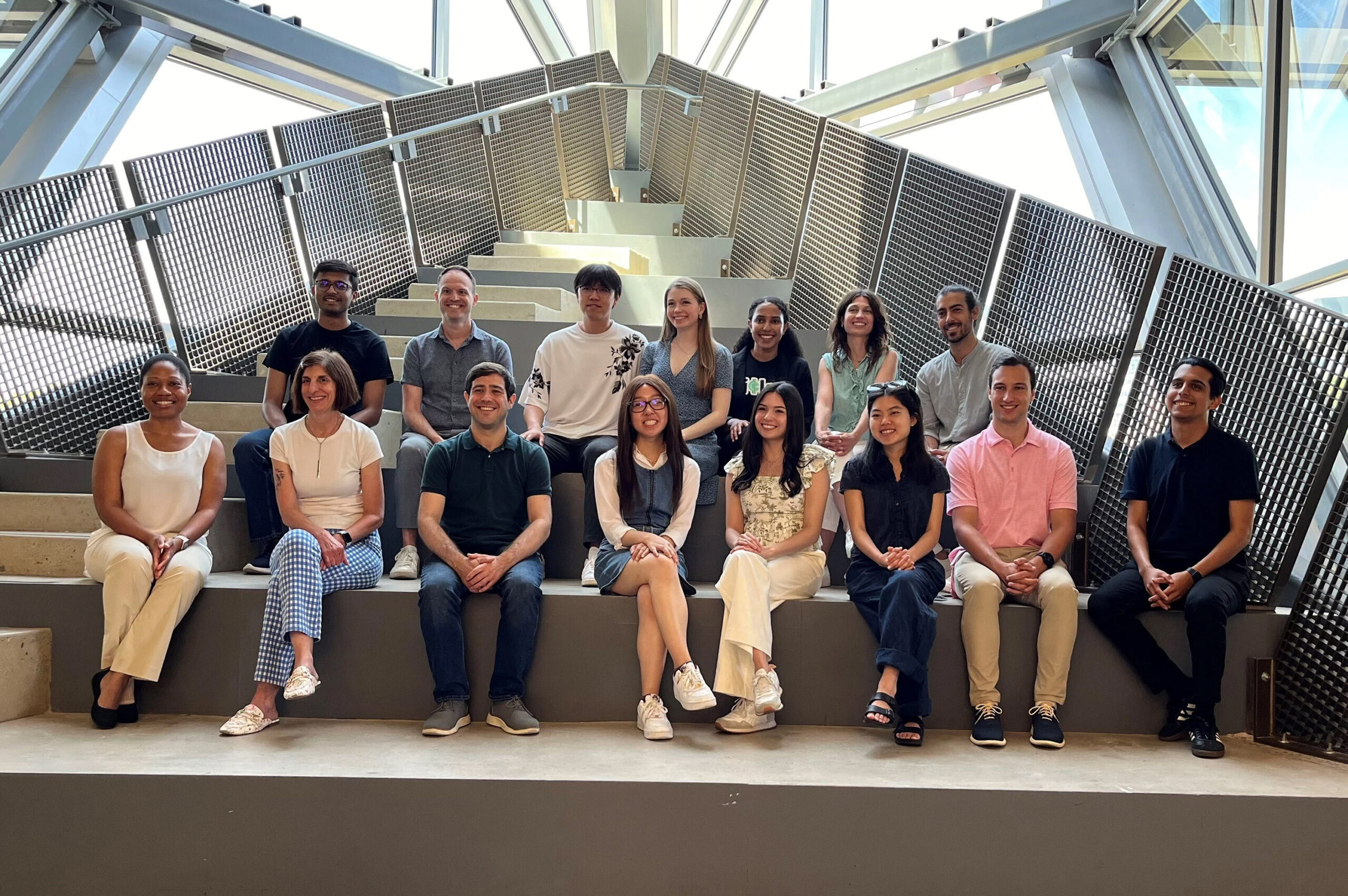 A group of fifteen people posing on a staircase inside a modern building with metal railings and large windows.