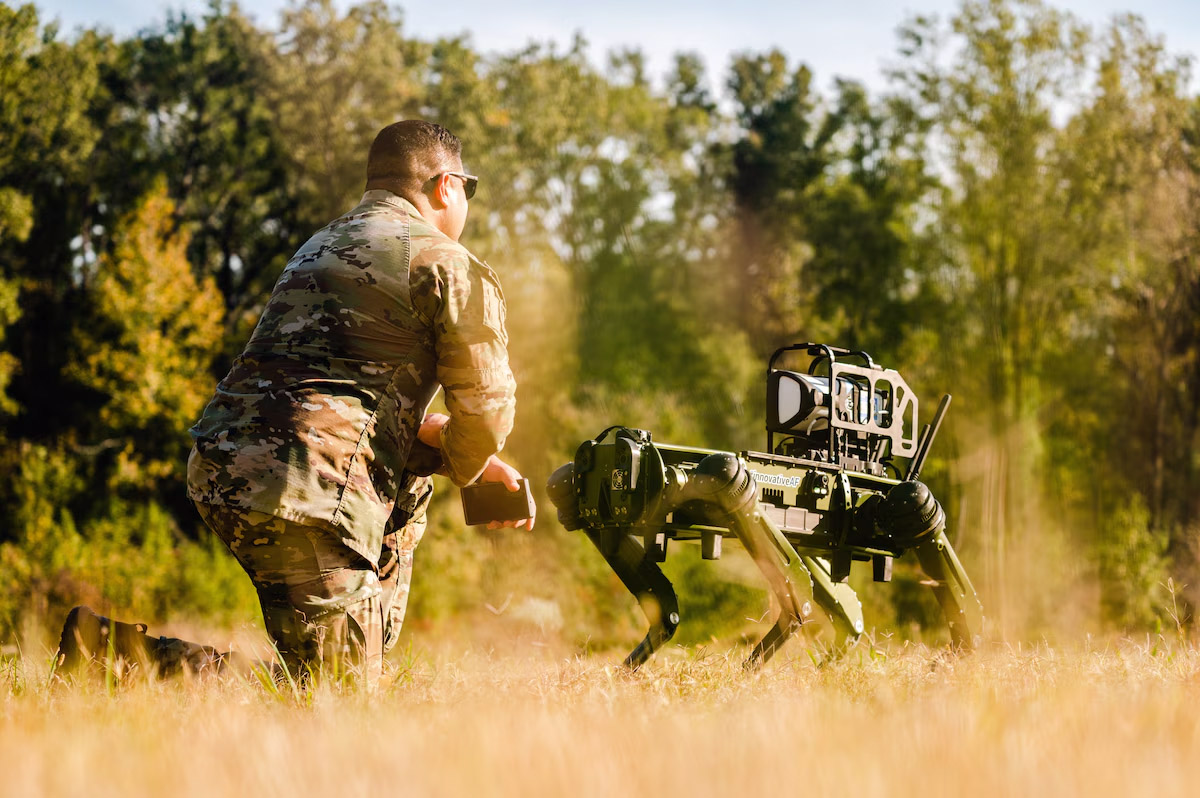 A person in military uniform interacts with a robotic quadruped in an outdoor setting with tall grass and trees in the background.