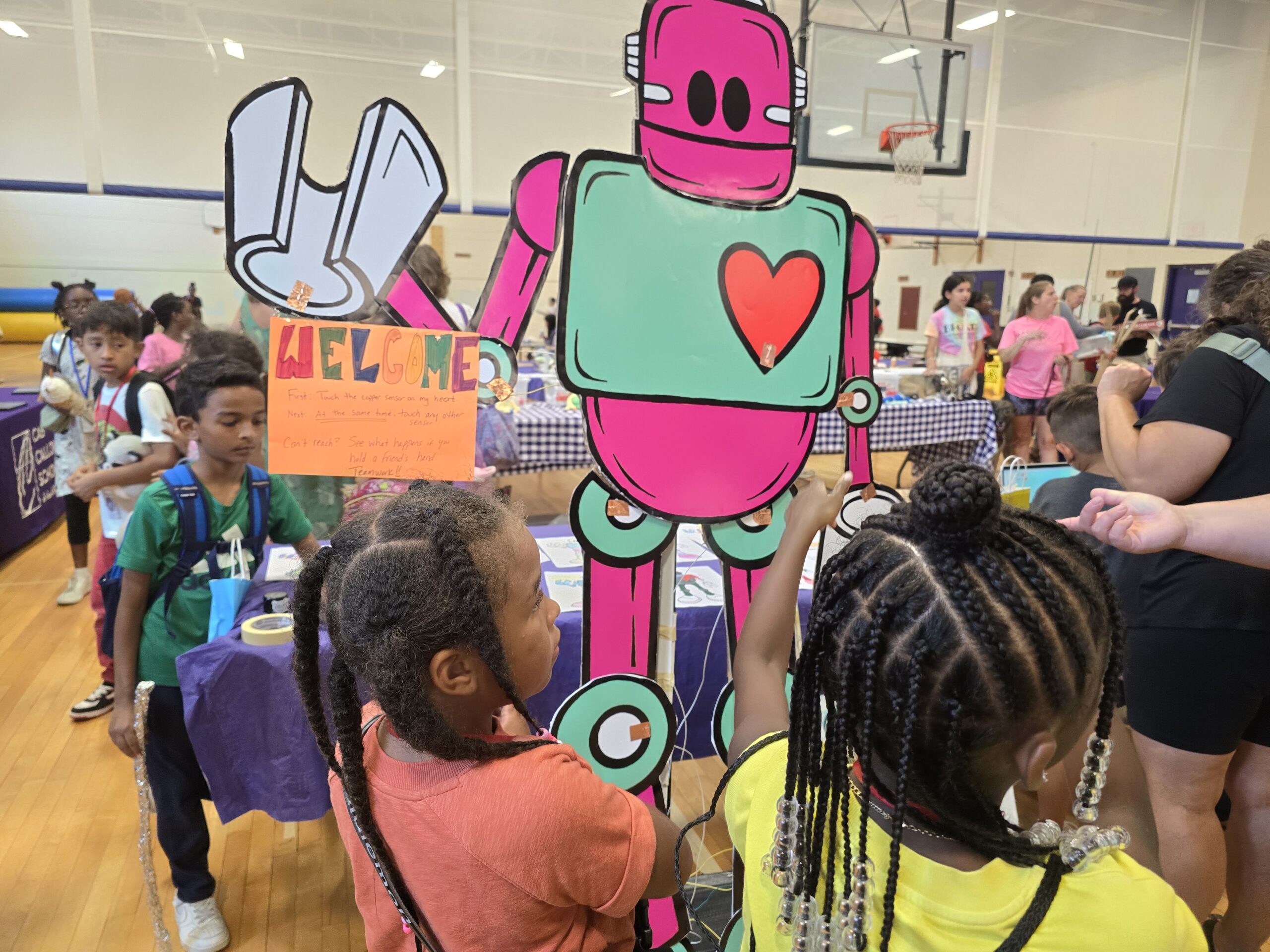 Children interact with a large pink robot cutout holding a "Welcome" sign at a school event in a gymnasium filled with various activity stations and other participants.