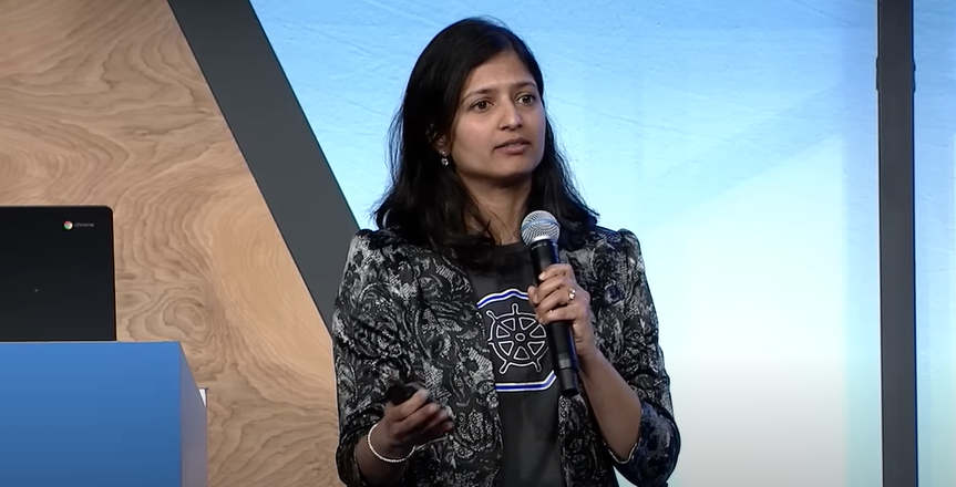 A South Asian woman with black hair holds a microphone in front of a brown and blue background