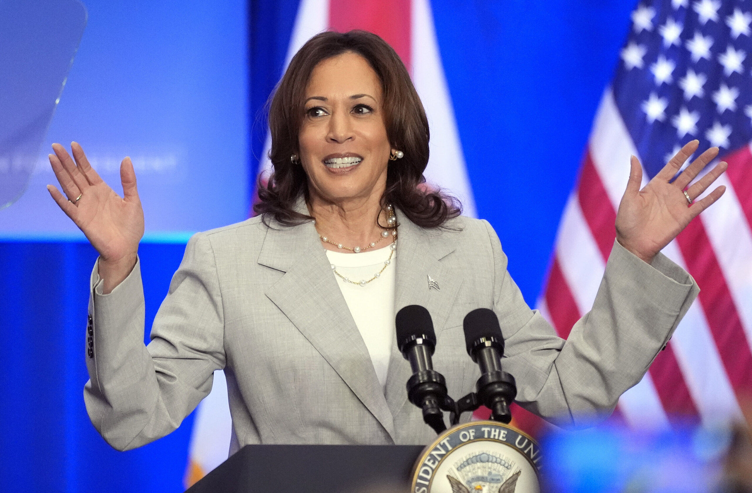 A woman in a gray blazer speaks at a podium with microphones, smiling, with flags in the background.