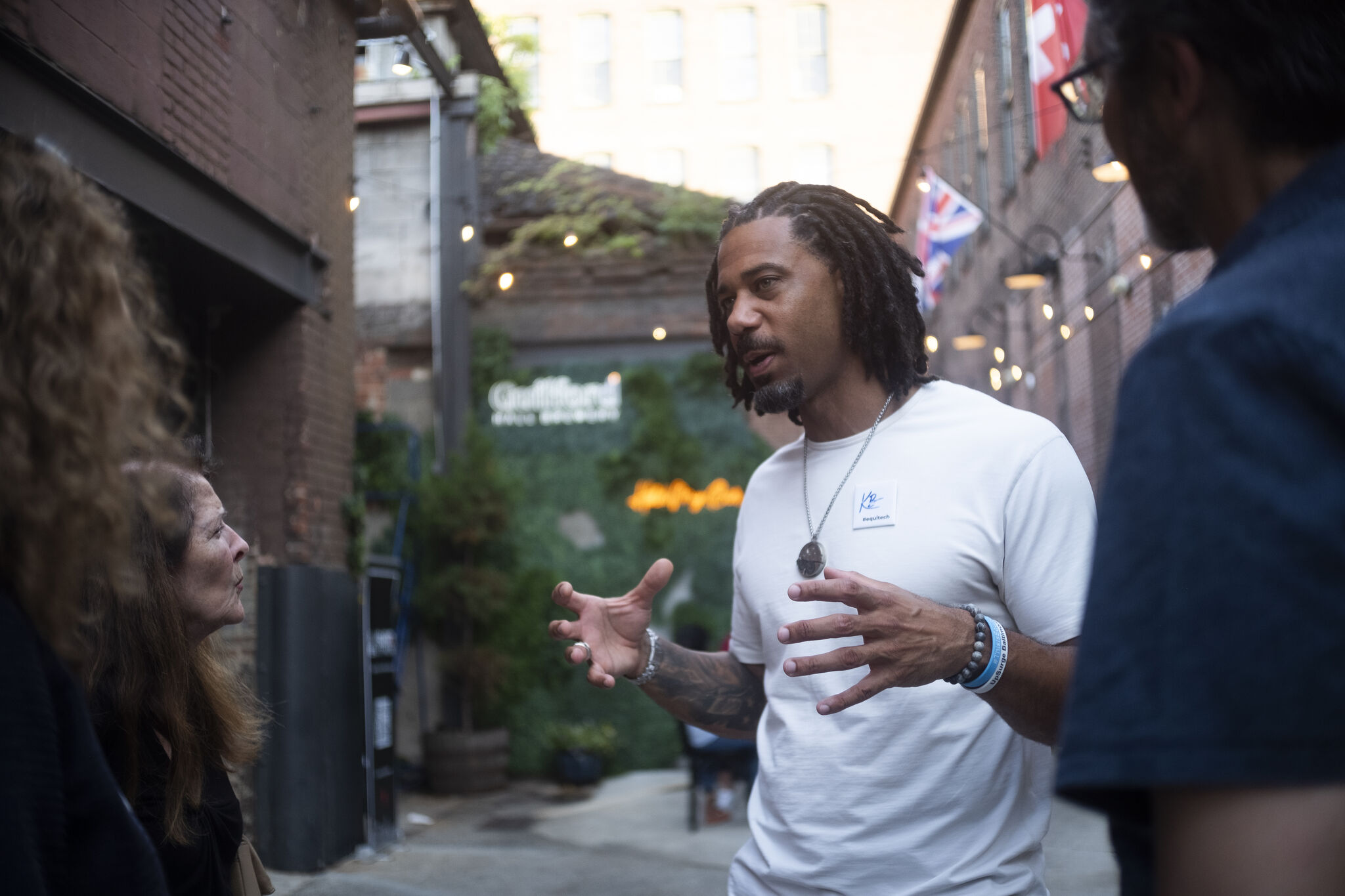 A Black man speaks to three people in an alleyway decorated with string lights and greenery.