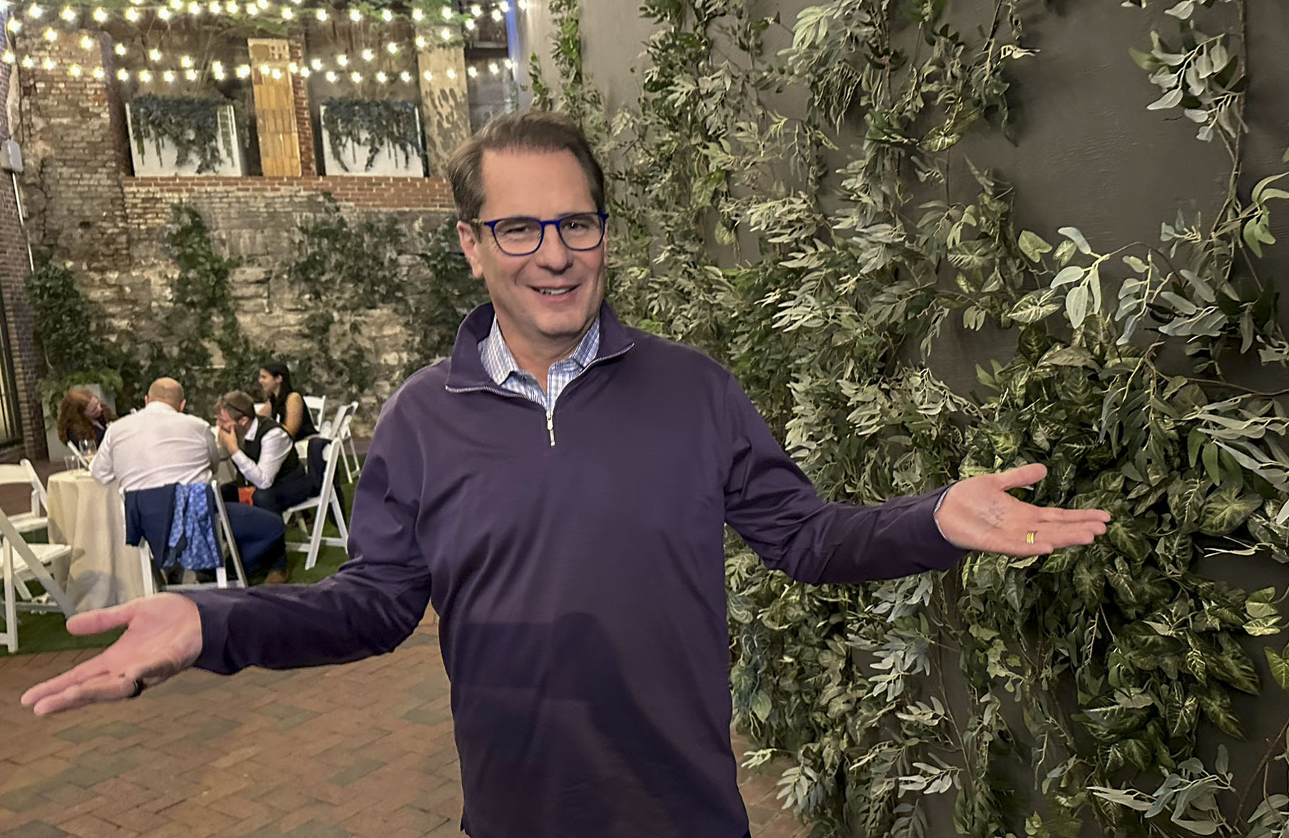 A white man smiling with arms outstretched in a courtyard decorated with hanging lights and greenery. People are seated at a table in the background.
