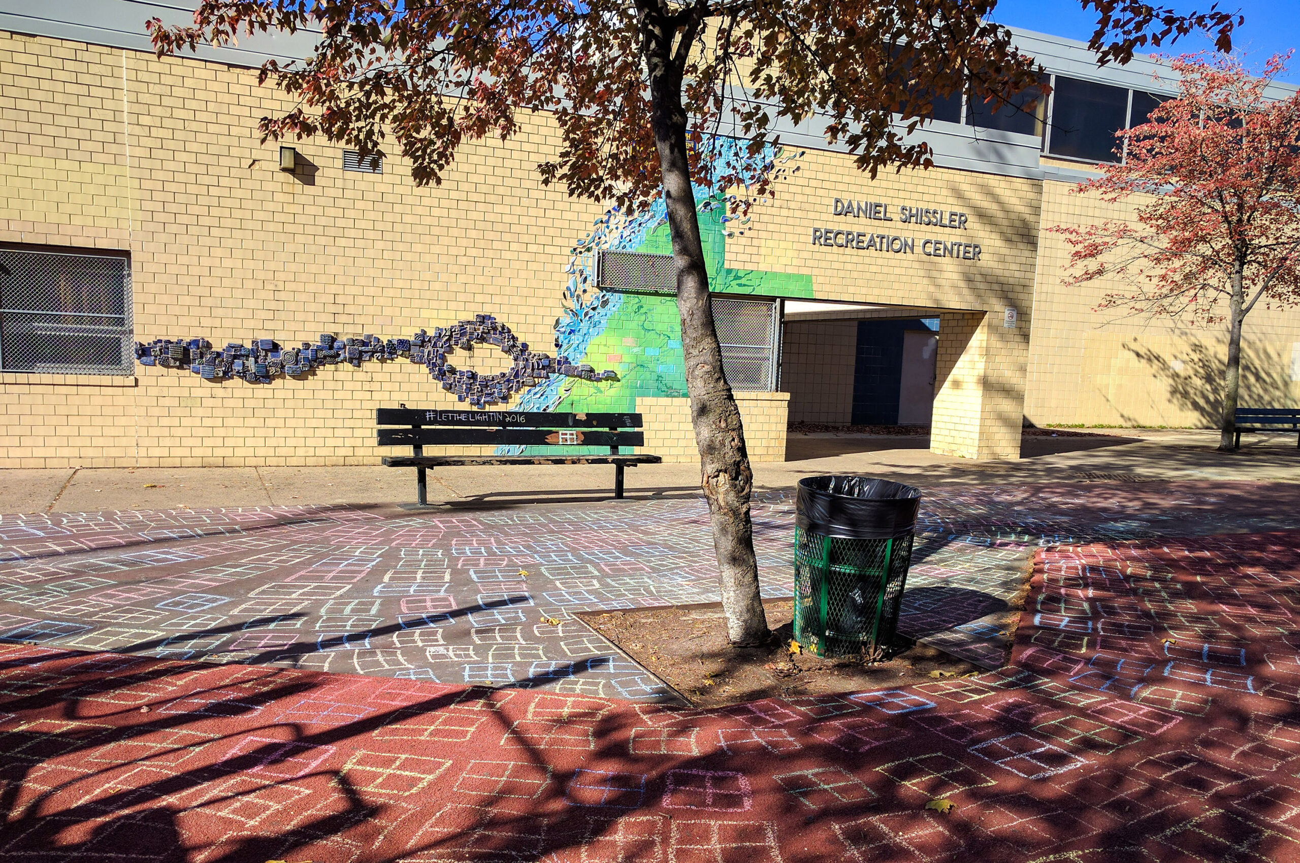 A brick recreation center with a wall mural, two trees, a black bench, and a black trash can. The ground is decorated with colorful chalk drawings.
