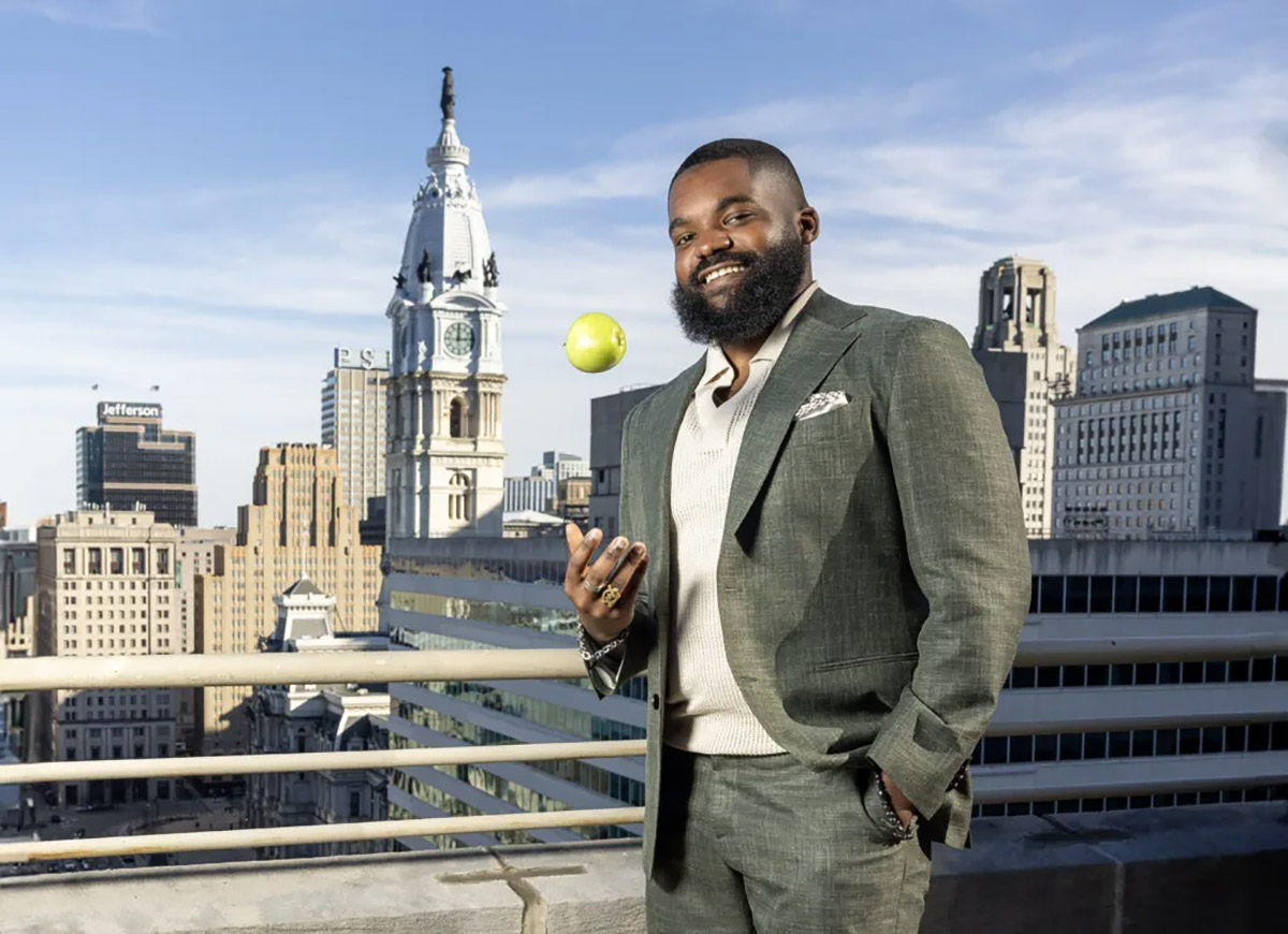 Man in a suit stands on a rooftop with cityscape background, smiling and tossing a green apple in the air.
