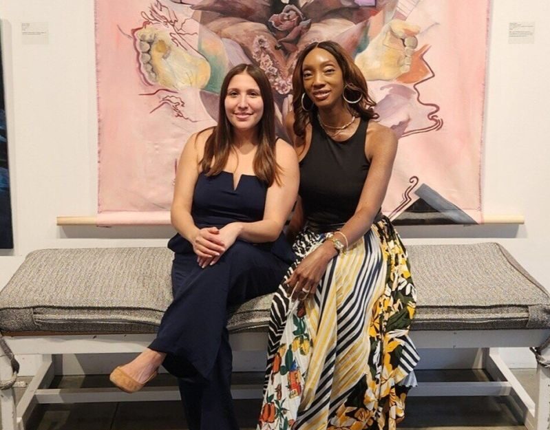 Two women sit on a bench in front of an art piece depicting multiple self-portraits of a person taking selfies. One woman wears a navy jumpsuit, and the other wears a black top and a colorful skirt.