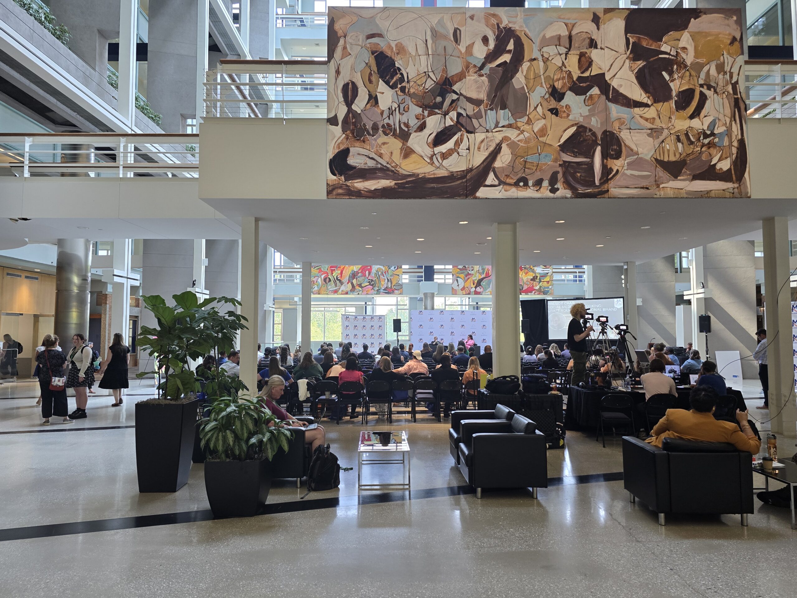 A modern, open-plan atrium with people seated facing a stage at the far end. Above the seating area is a large, abstract mural. There are tall plants and sofas in the foreground.