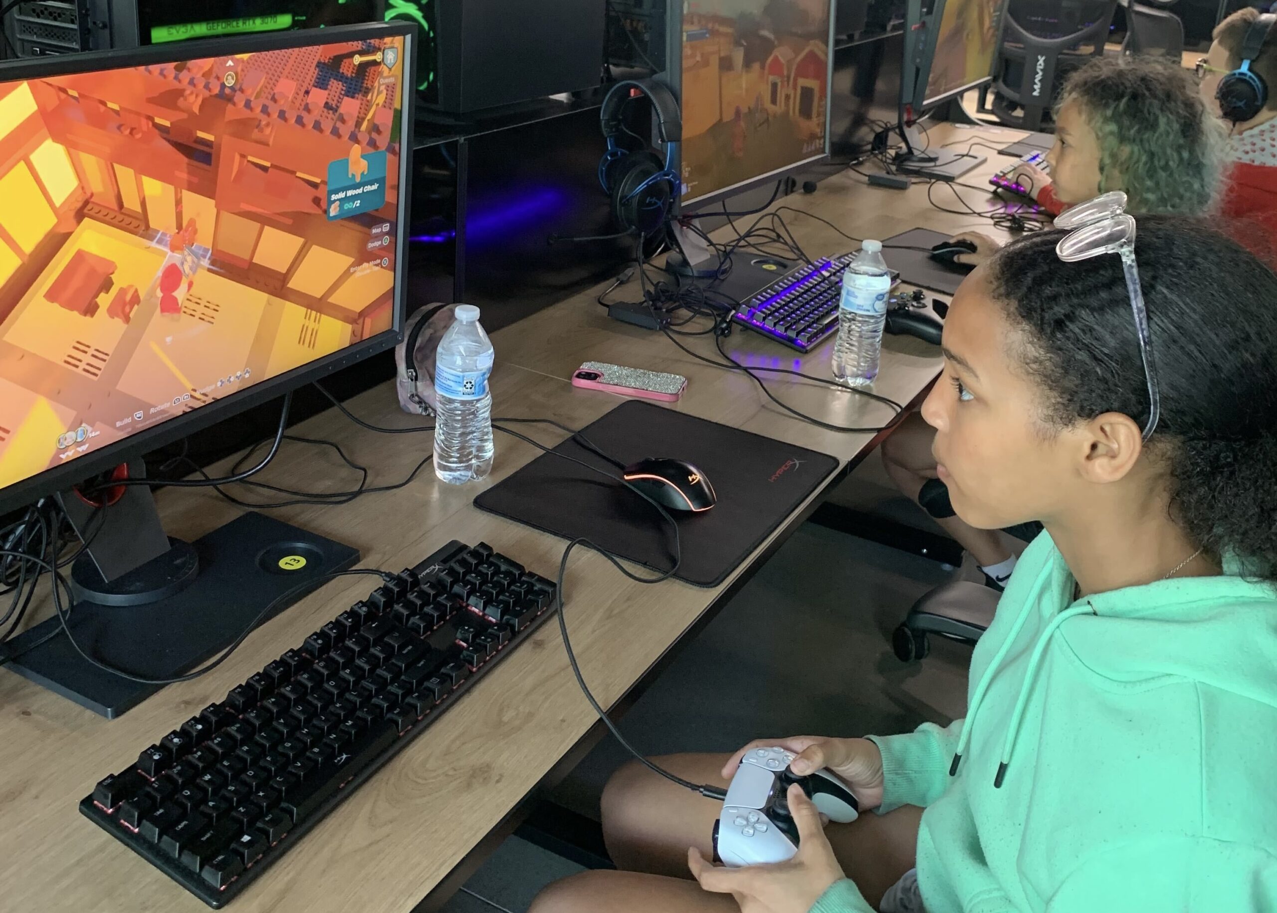 A person in a green hoodie is sitting at a desk with a PlayStation controller, playing a game on a computer monitor. Other people are seated at adjacent desks, each with similar setups.