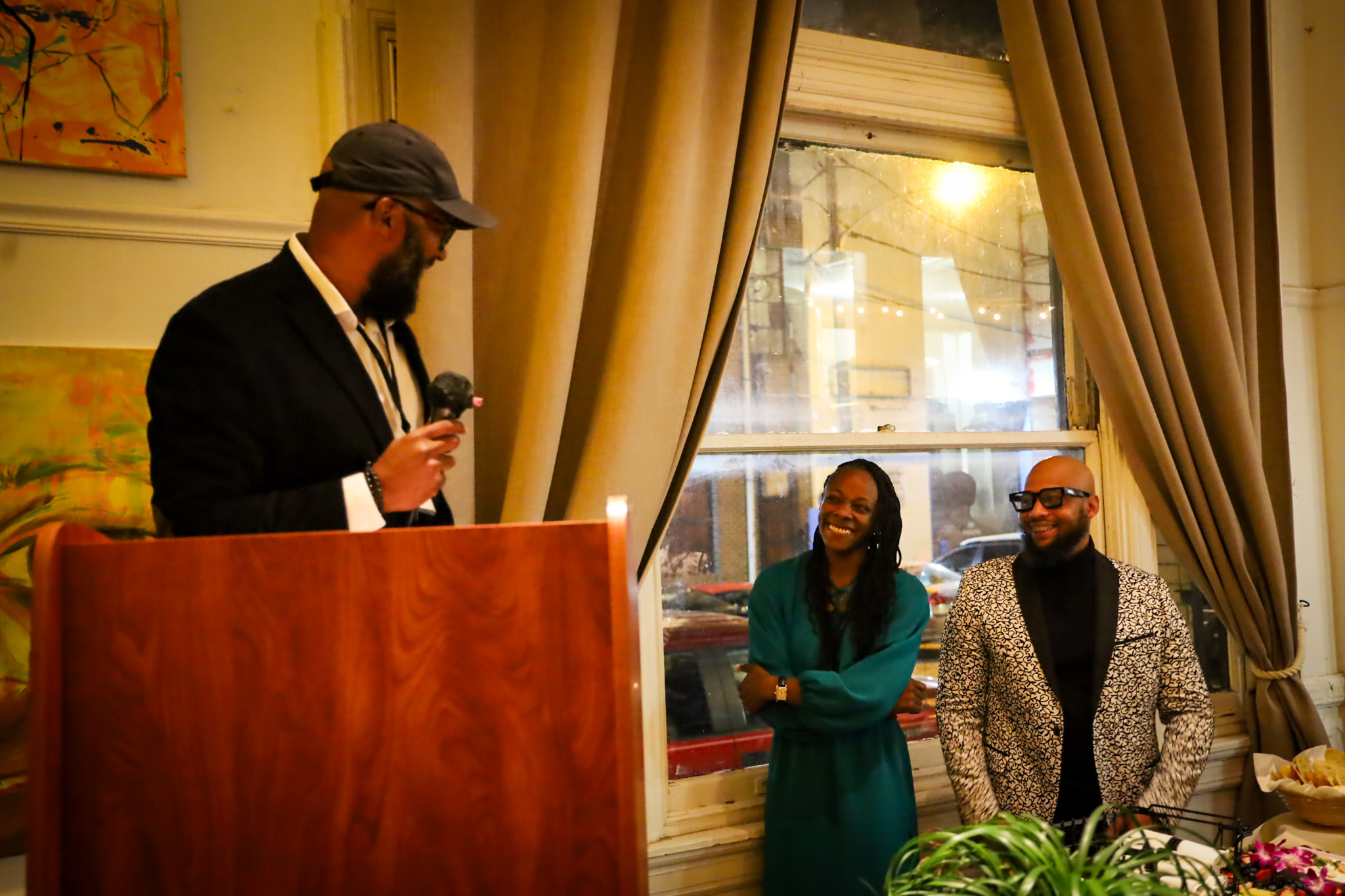 A person at a podium speaks into a microphone while two people stand near a window smiling and listening.