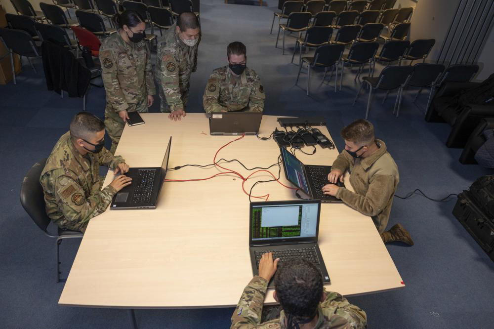 People in camo fatigues stand and sit around brown table with black laptops. to do things