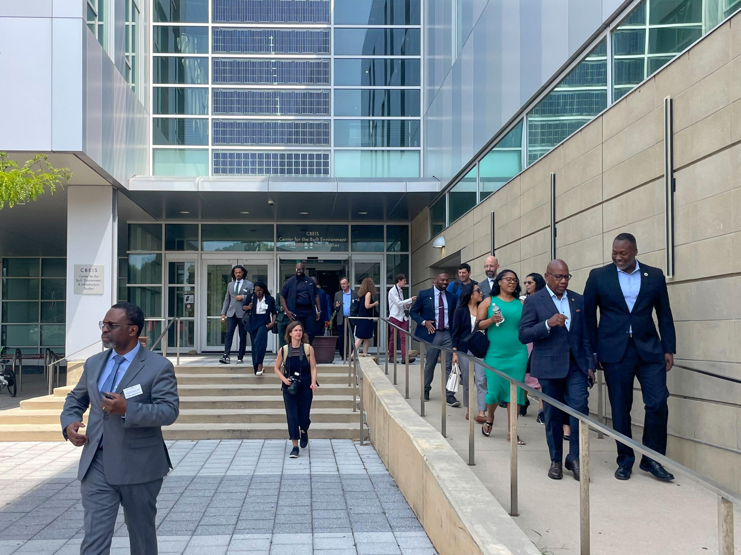 A group of people in formal attire walk down the steps and ramp outside a modern glass building.