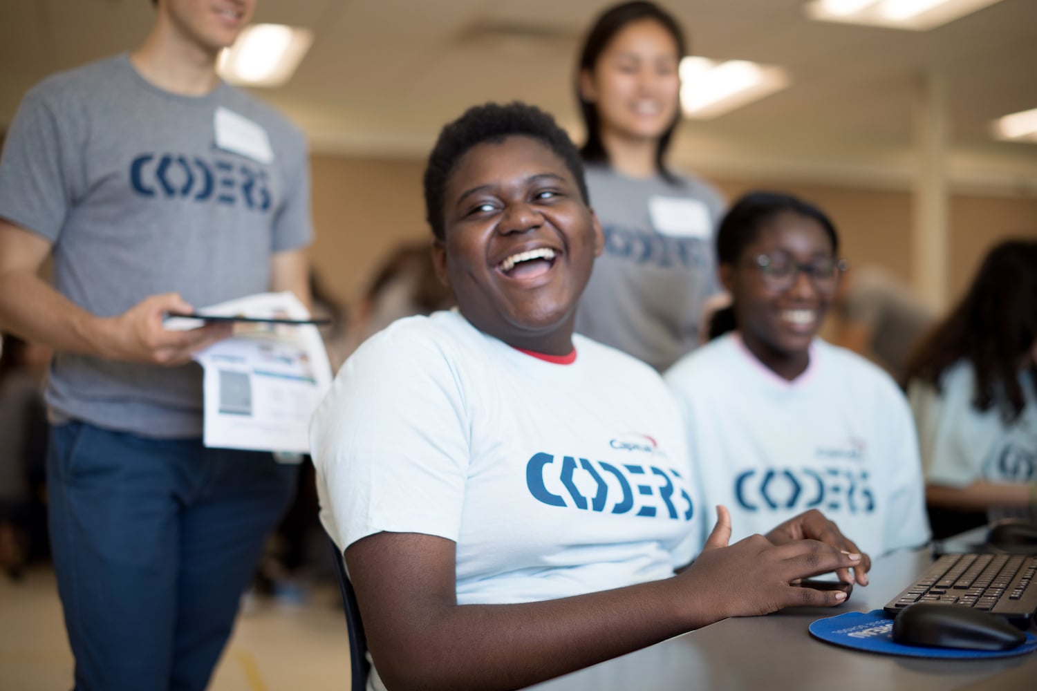 A group of young participants, wearing "Coders" t-shirts, smiling and engaged in front of computers during a coding event.