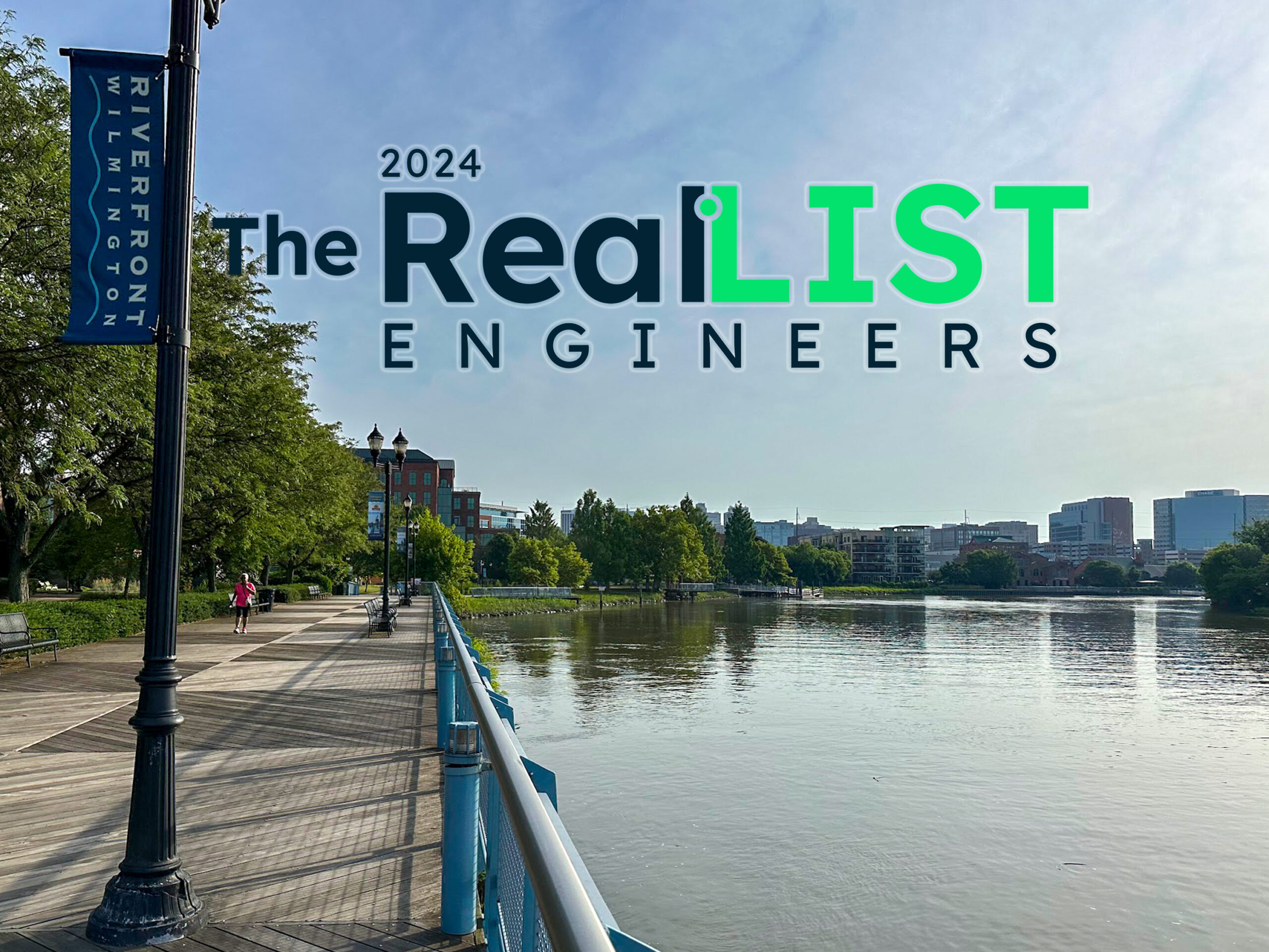A riverfront boardwalk in Wilmington with trees, buildings in the background, and a large banner on the boardwalk railing. Text reads "2024 The Real List Engineers."