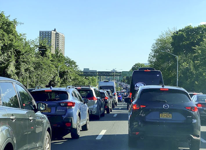 Heavy traffic on a multi-lane road with numerous cars and a bus stuck in congestion, greenery on both sides, and a tall building visible in the background under a clear blue sky.