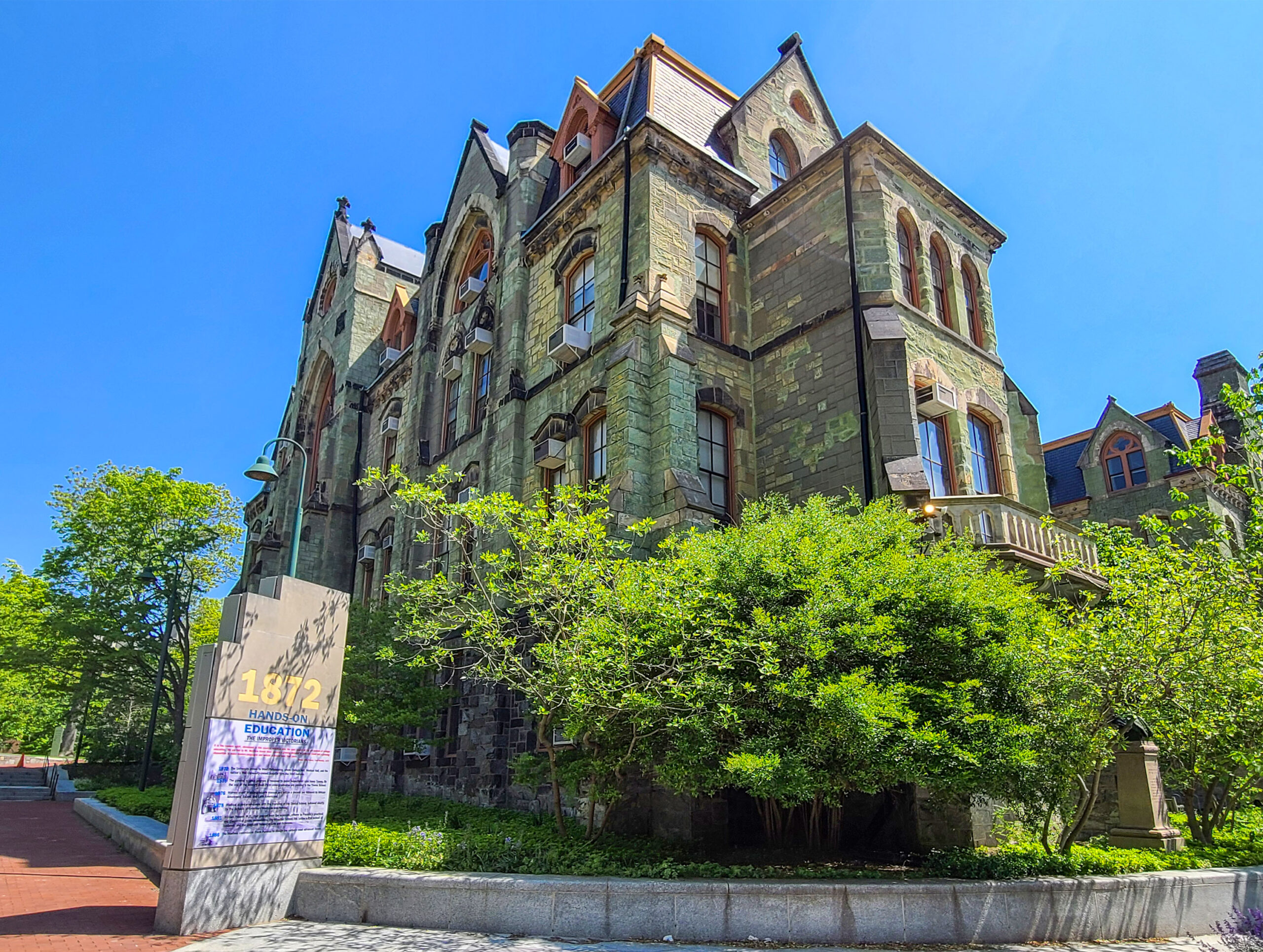A stone building with tall, pointed roofs, arched windows, and green foliage around it under a clear blue sky. A sign dated 1872 stands in the foreground.