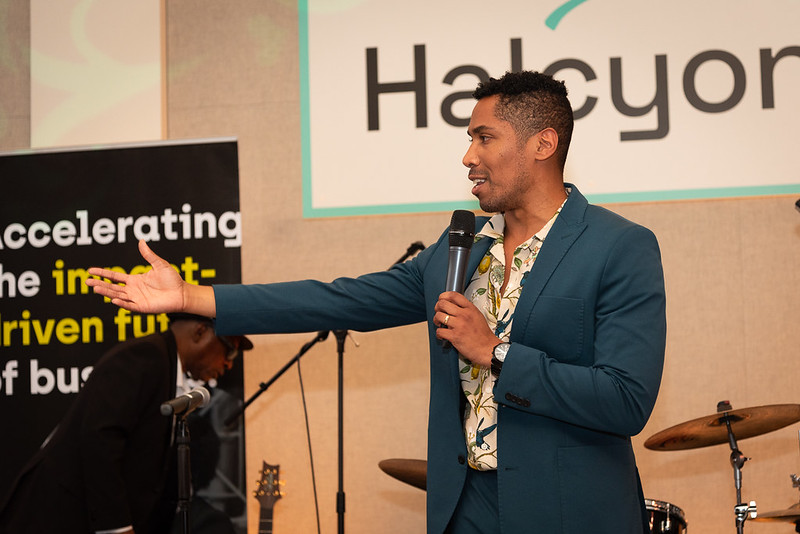 A man in a suit speaks into a microphone on stage, gesturing with one hand. A drum set and a sign reading "Halcyon" are visible in the background.