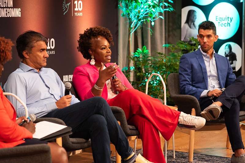 A woman in a pink blazer speaks while seated on a panel with two men. The panel discusses equity in tech, as indicated by the backdrop.