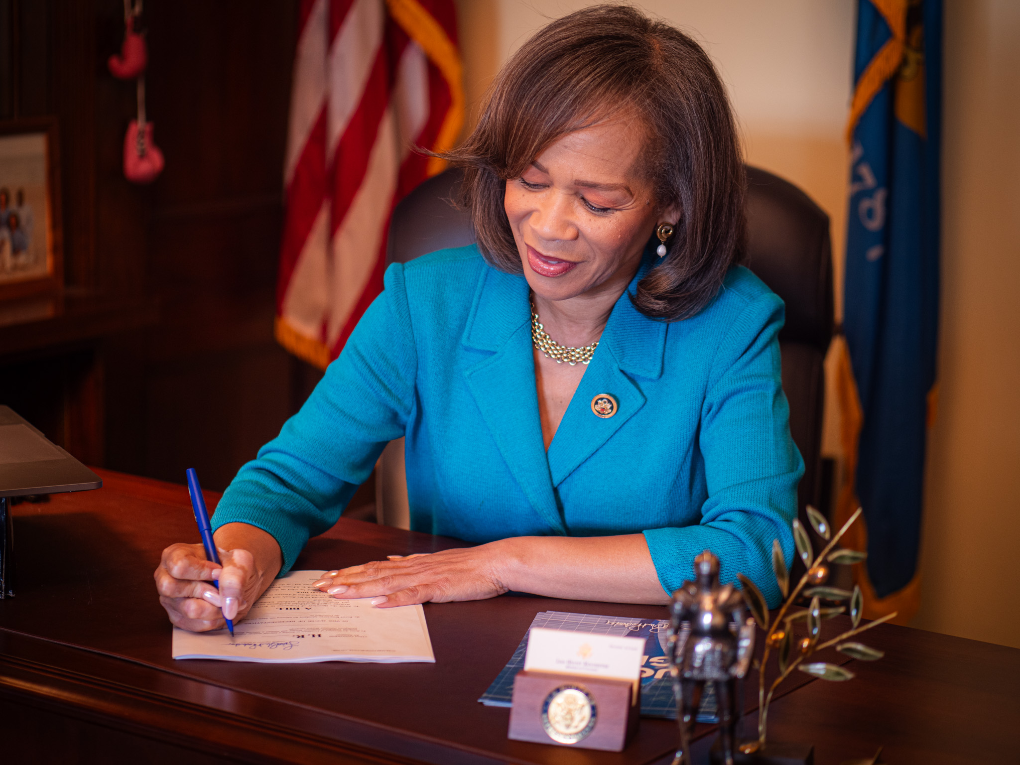 A woman in a blue blazer signs a document at a desk, with a small statue, nameplate, and flag in the background.