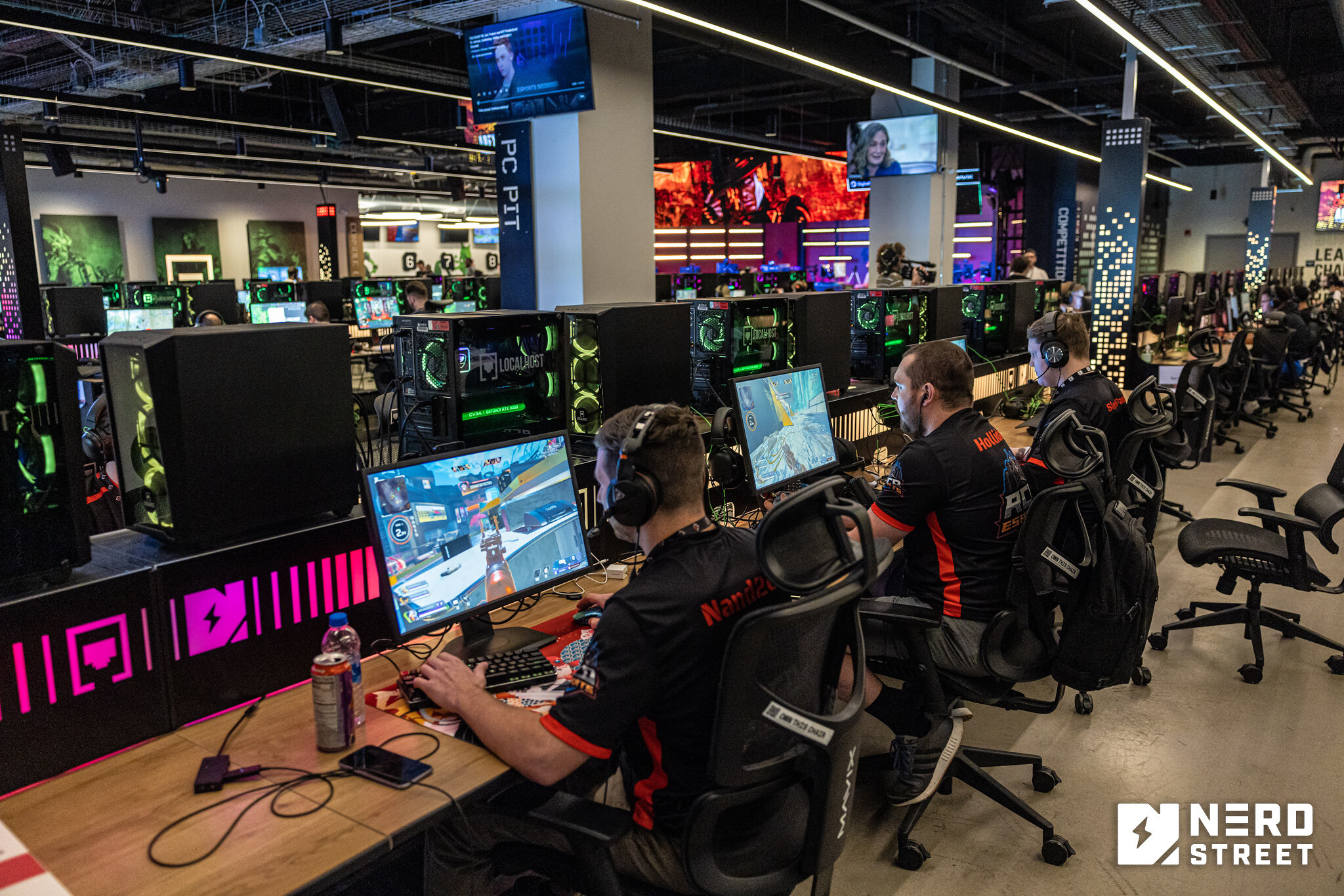 Gamers wearing headsets and jerseys compete in an esports tournament at a gaming facility equipped with high-end PCs. The facility has rows of computer stations and colorful lighting.