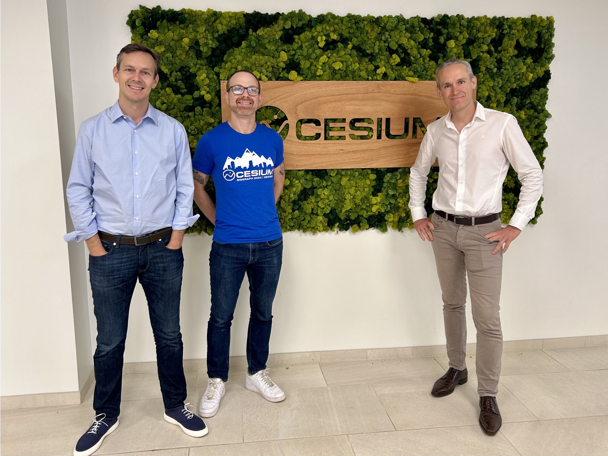 Three men standing in front of a wall with a "Cesium" sign, two in casual attire and one in business attire. The backdrop features a leafy green design with a wooden sign.