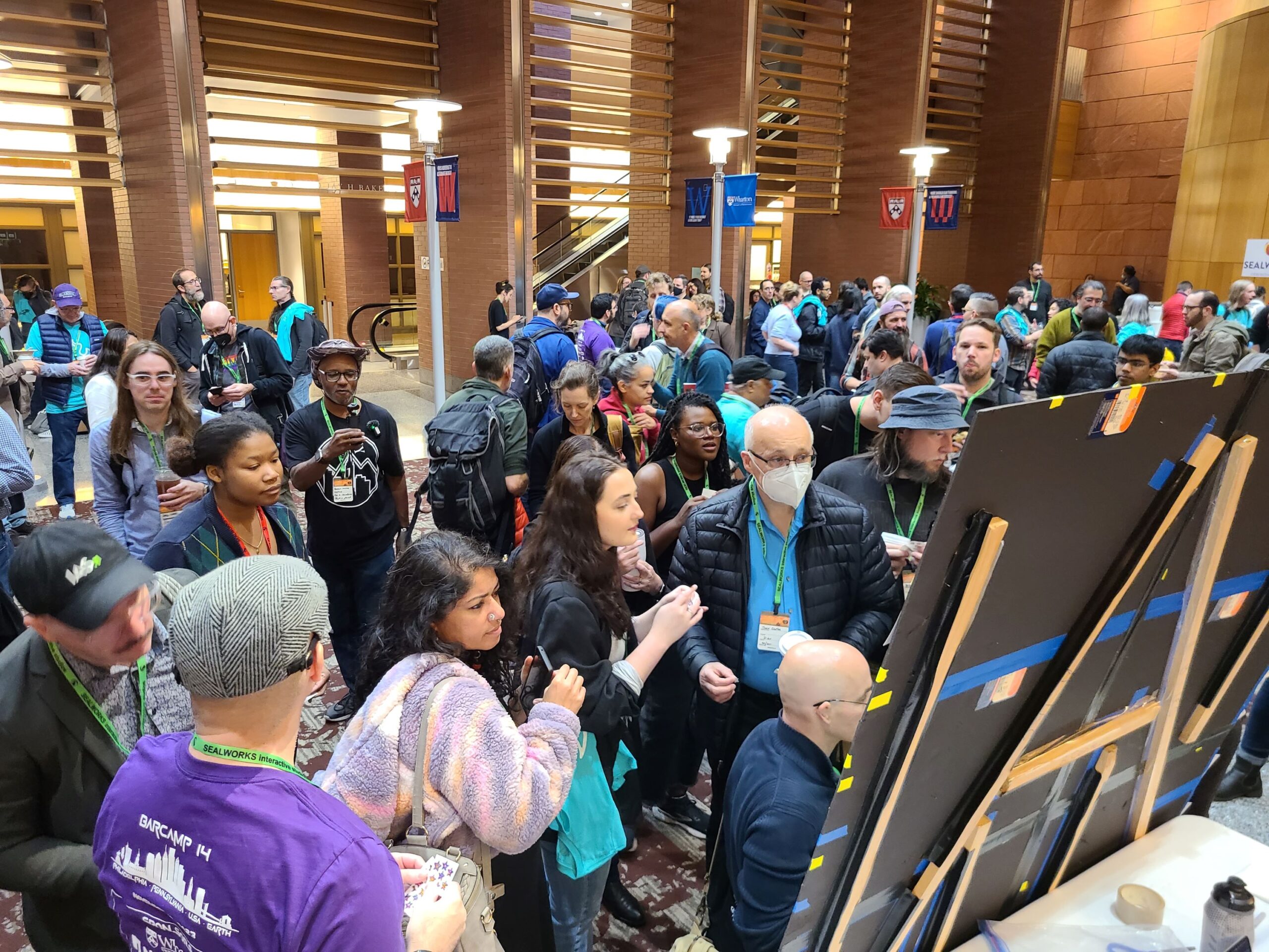 People gathered in a lobby area near a large board, some looking at the board and others engaged in conversation. The space features modern wooden architectural elements and banners.
