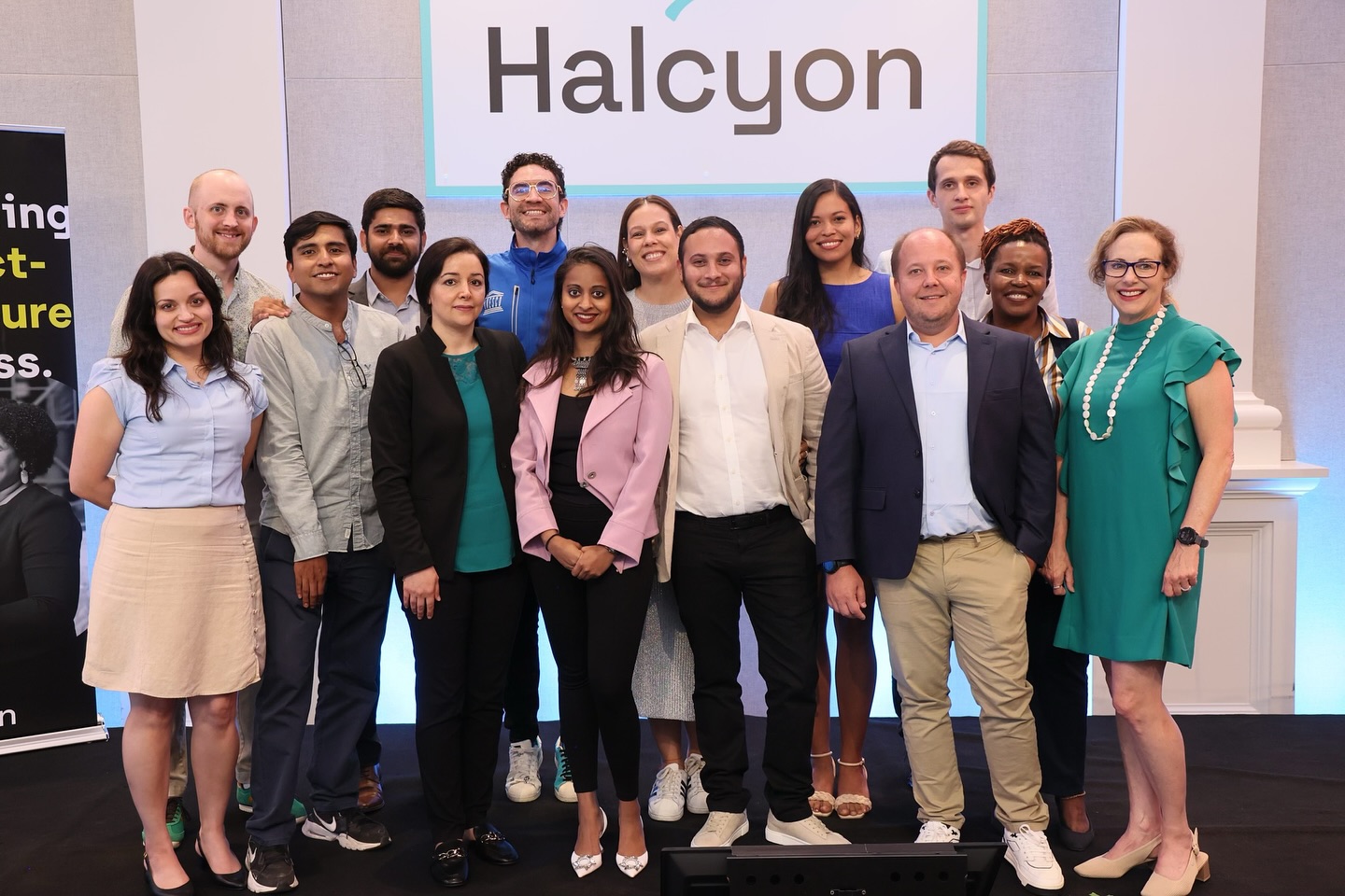 A group of people stands and poses for a photo in front of a "Halcyon" sign, featuring a mix of men and women dressed in smart casual to business attire.