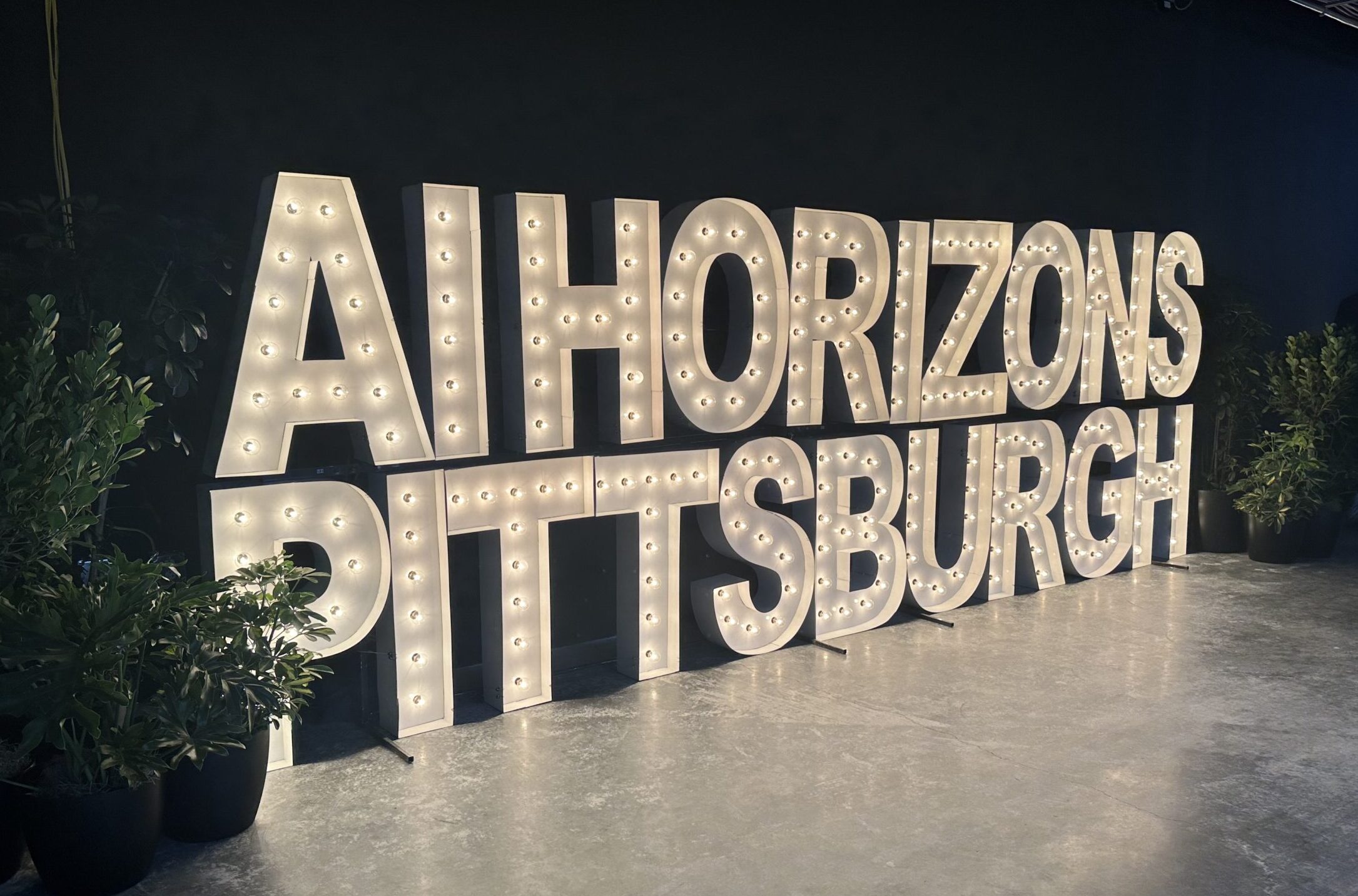 Lit marquee letters spell "AI HORIZONS PITTSBURGH" against a dark background with plants on either side.