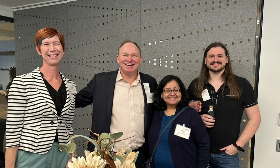 Four people are standing indoors, posing together for a photo. They are smiling and dressed in business casual attire, with conference name tags visible.