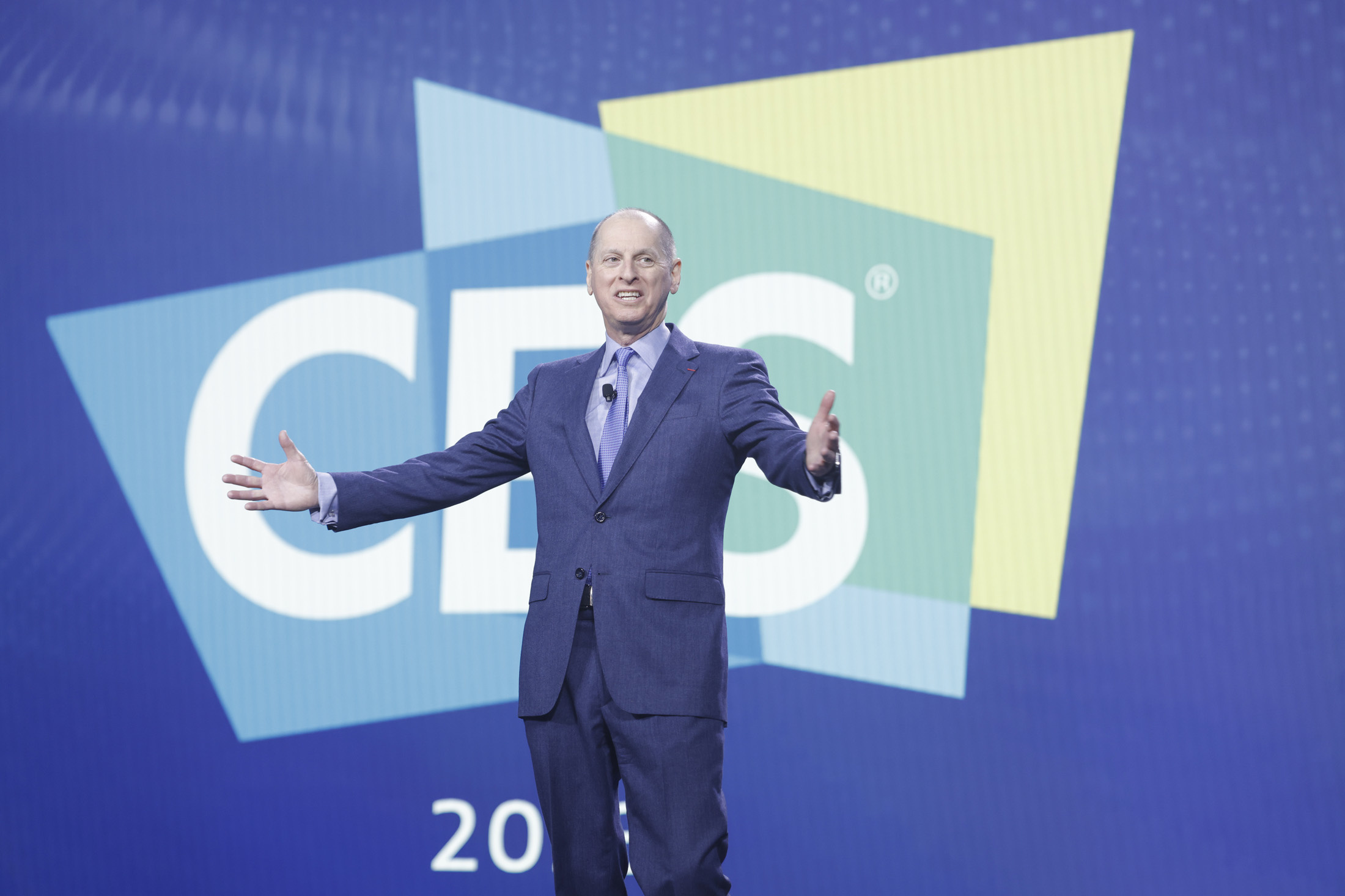A bald white man in a blue suit holds his hands out in front of a blue wall with multicolored logo