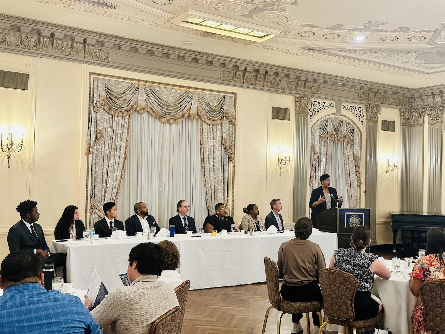 A panel of nine people sits at a long table during a formal event in an ornate room. An individual stands at a podium speaking to an audience. Attendees are seated at round tables in the foreground.
