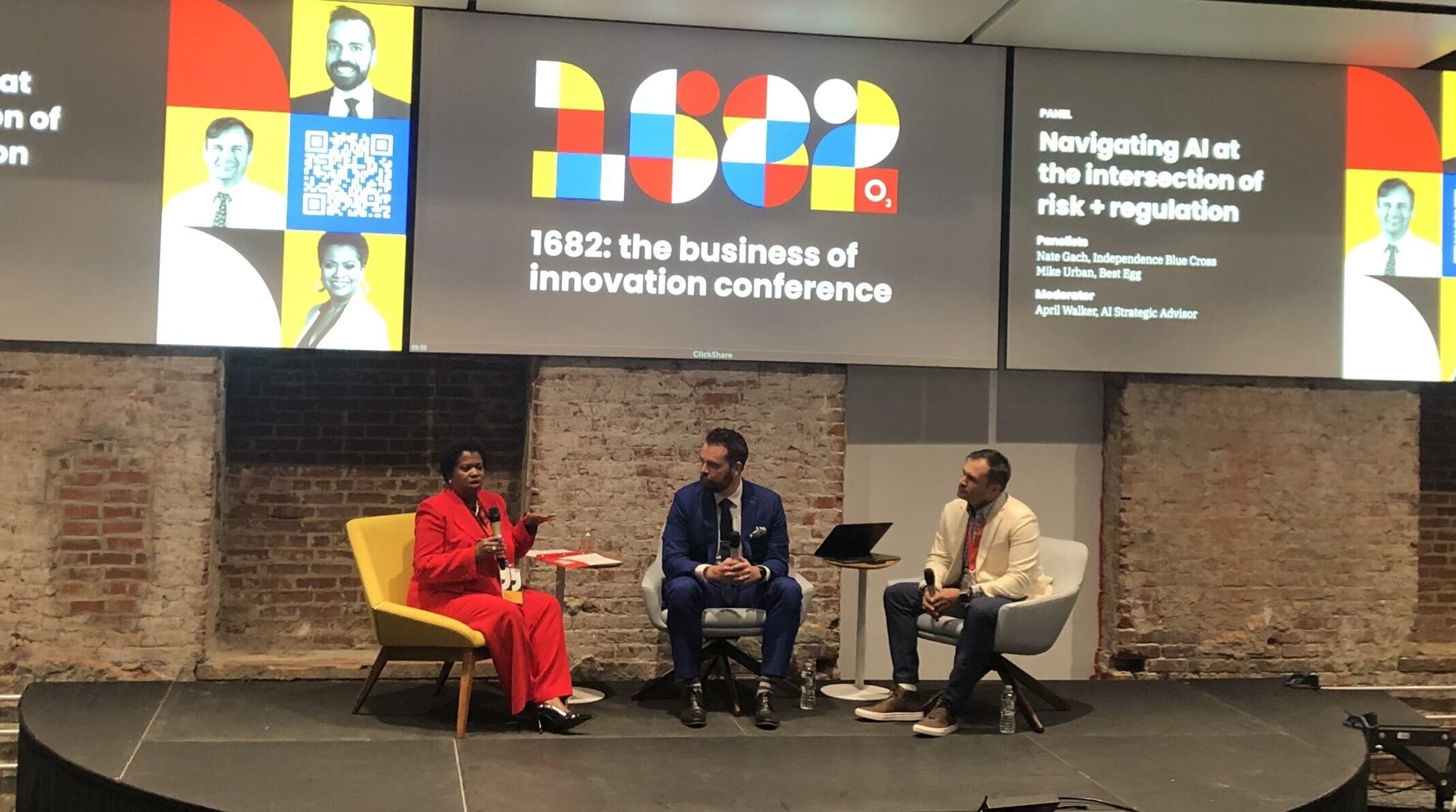 Three people sit on a stage in a panel discussion at the "1682: The Business of Innovation Conference." Two large screens display event details above the participants.