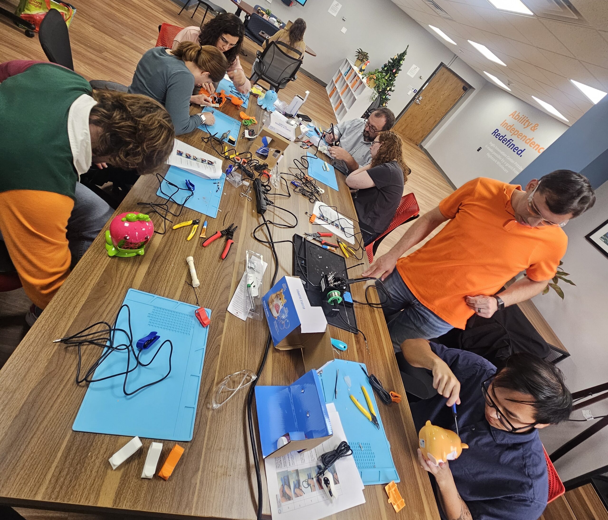 A group of people are sitting and standing around a table working on various projects with electronic components and tools.
