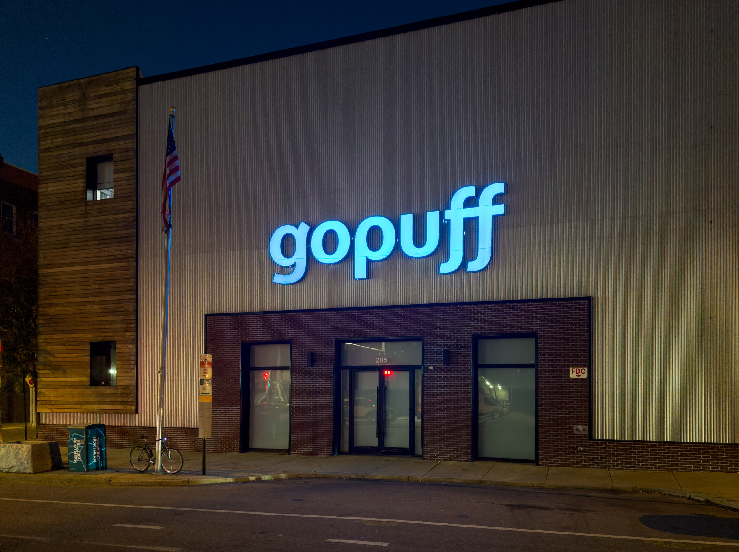 Nighttime exterior of a building with a "Gopuff" sign, a flagpole, and a bicycle parked by the entrance.