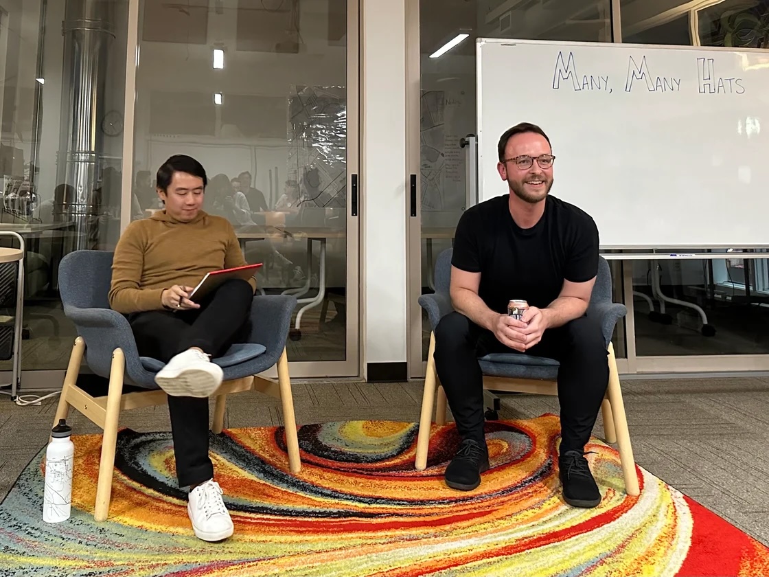 Two people seated in chairs in an office setting. One is holding a red tablet, the other has a can. A whiteboard in the background reads "Many, Many Hats.