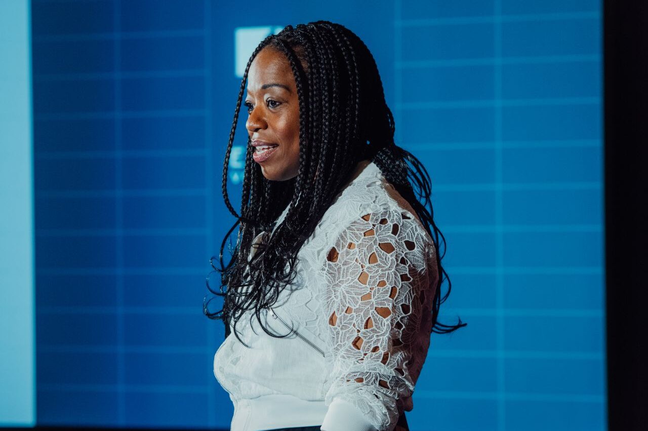 A person with long braided hair speaks in front of a blue screen. They are wearing a white, lace-detailed blouse.