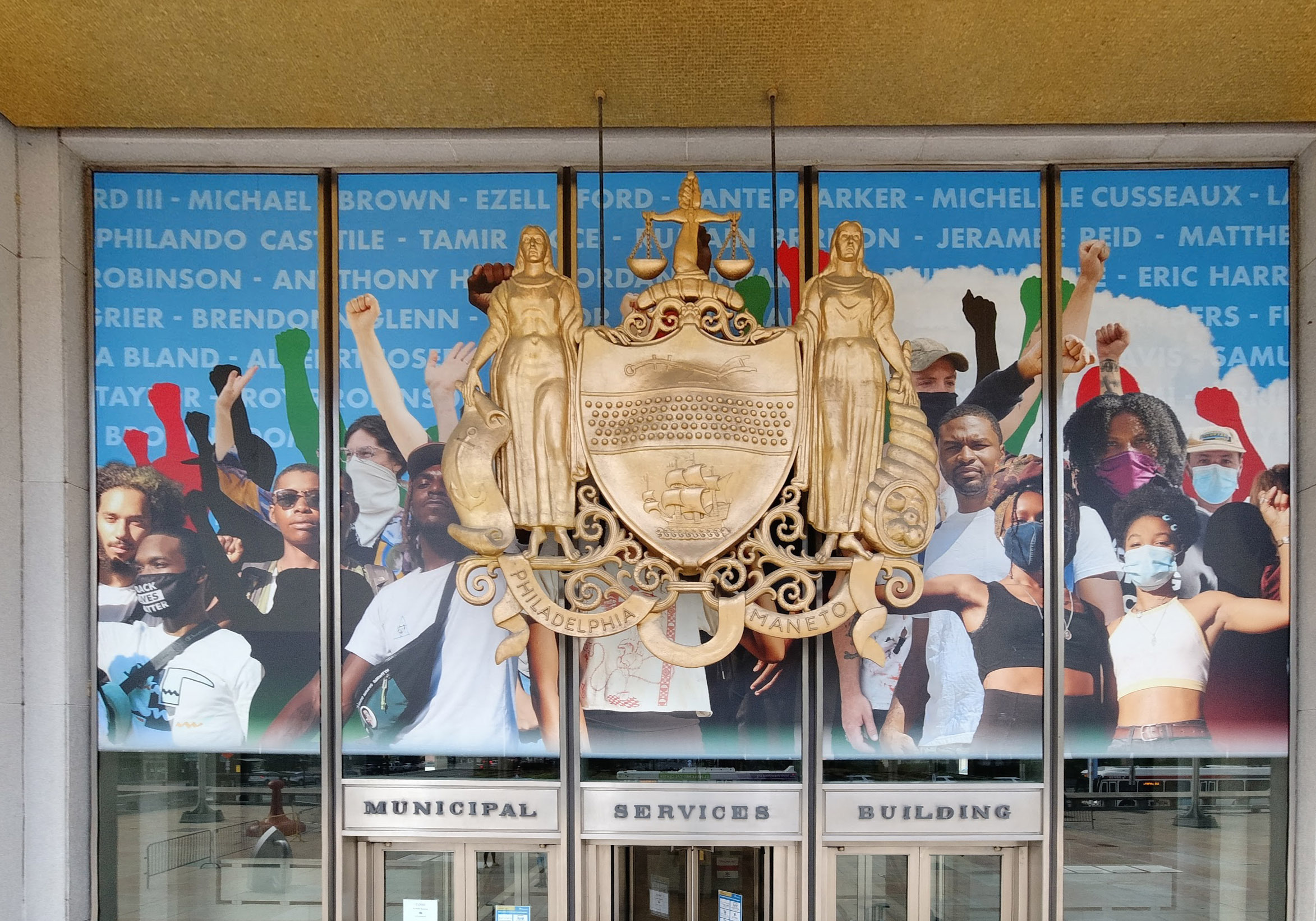 Entrance to a municipal building with a gold emblem and a mural featuring diverse individuals in protest, holding up fists.
