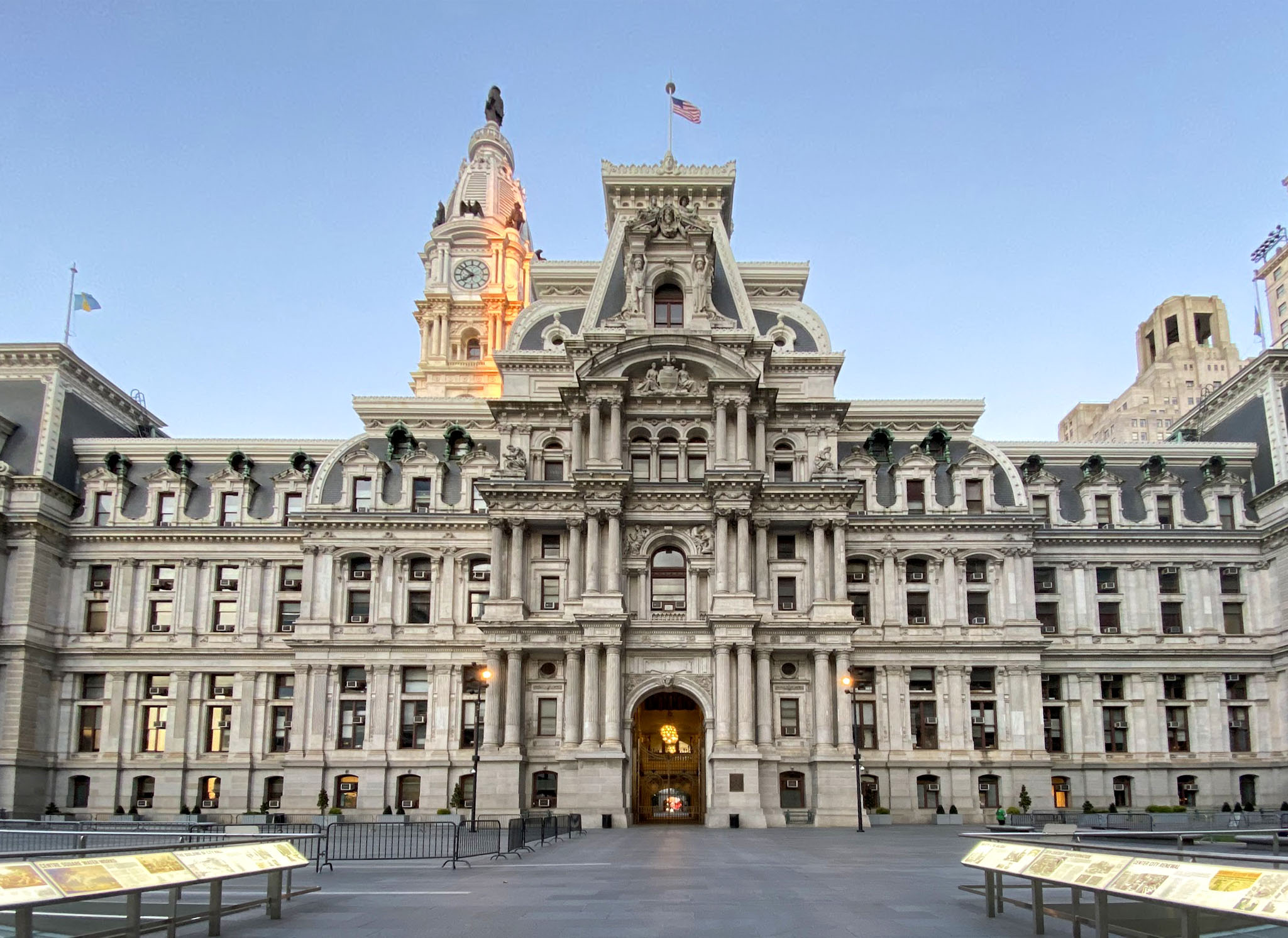 The image shows an ornate, historic building with a clock tower and statues. An American flag is flying on top. The facade has multiple windows and decorative elements.