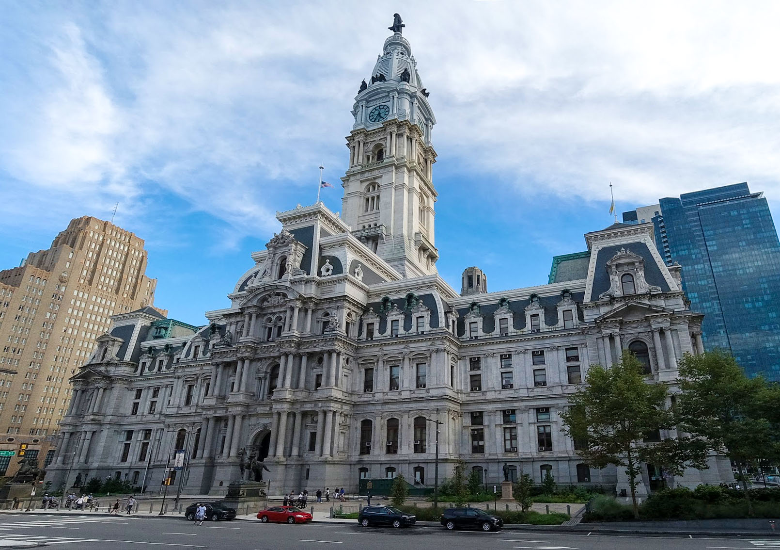 Historic building with ornate architecture, featuring a tall clock tower. Several people are walking nearby, and there are cars on the street in front. Skyscrapers are visible in the background.