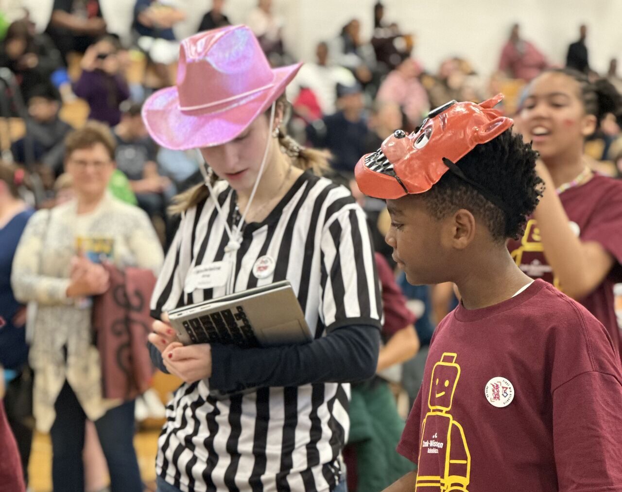 A boy in a maroon shirt participates in a robotics competition, observed by a person in a striped shirt with a notepad. A crowd watches in the background.